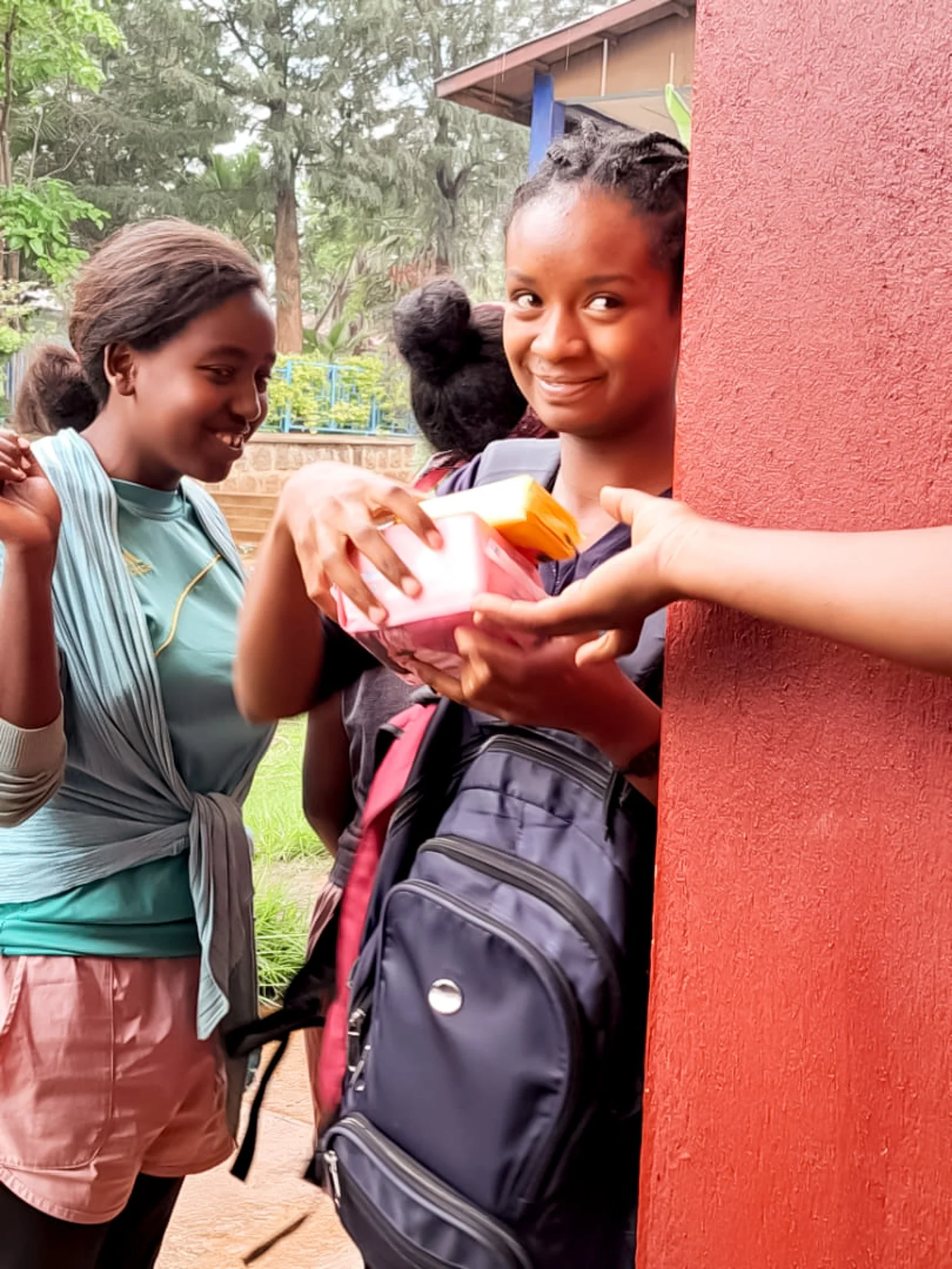 A young woman with braided hair is smiling as she receives a pink gift bag from someone whose arm is visible, partially hidden behind a red wall, during daytime outdoors.