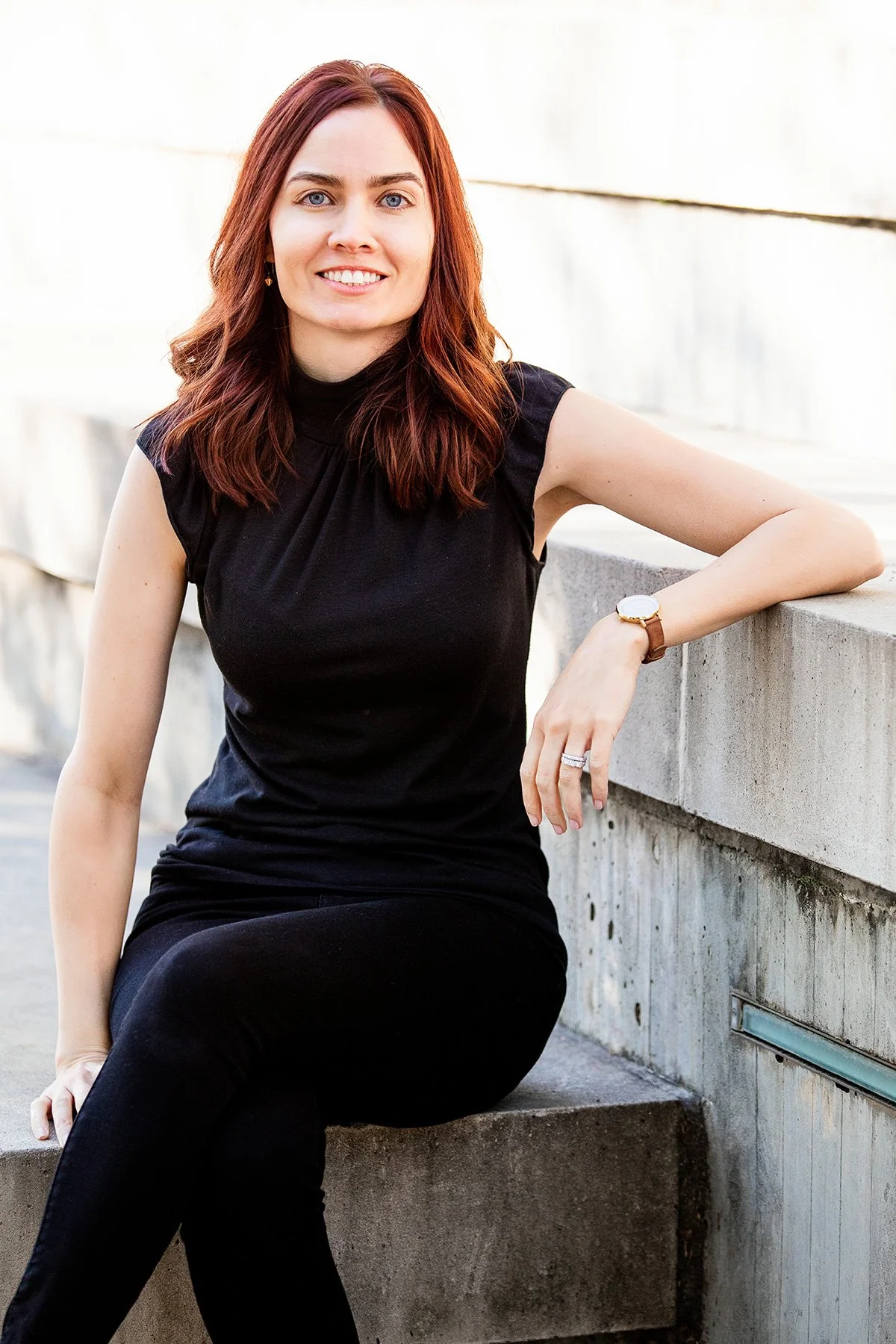 A woman with shoulder-length auburn hair and blue eyes, wearing a sleeveless black shirt and black pants, sitting outdoors beside a concrete wall, smiling at the camera.