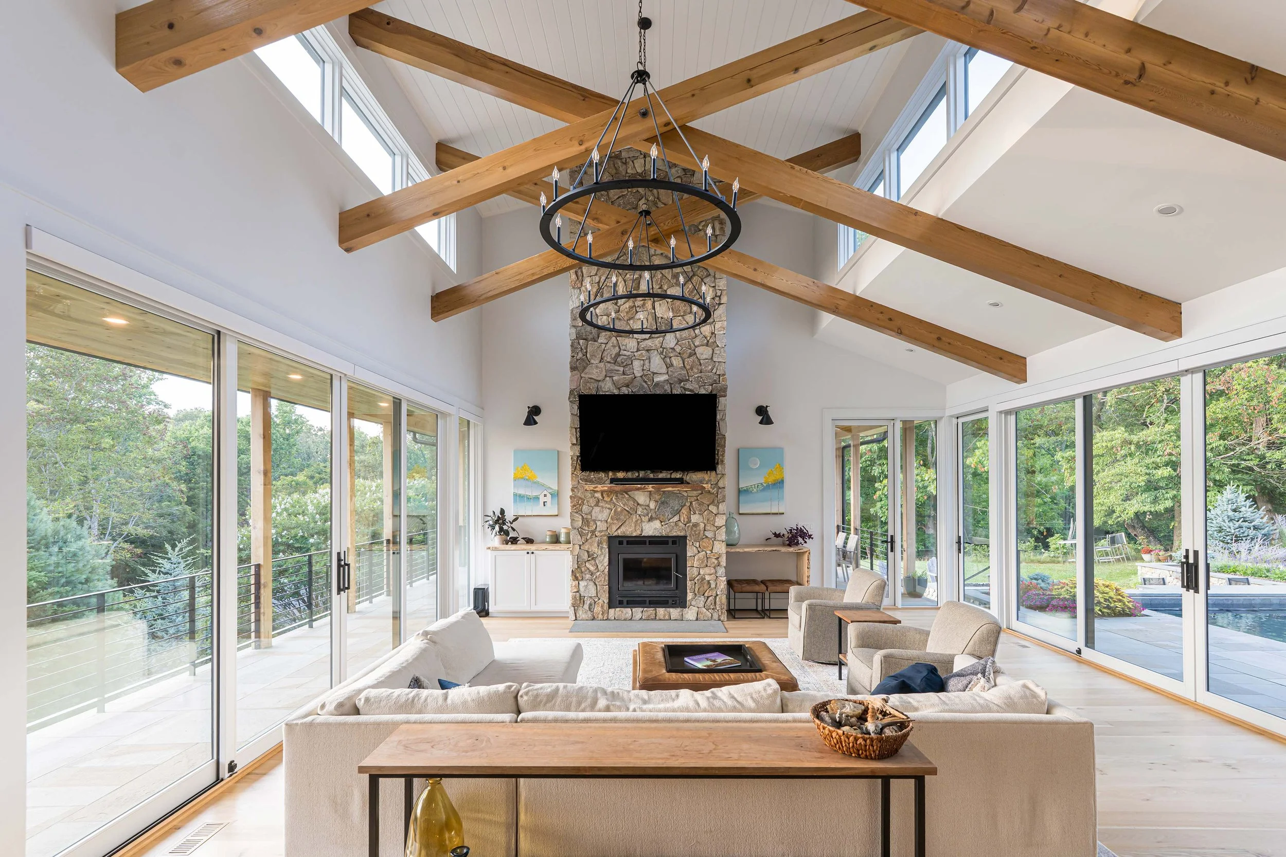 Bright living room with large glass sliding doors, high ceiling with exposed wooden beams, a stone fireplace with a mounted TV, and modern chairs and sofa facing the fireplace.