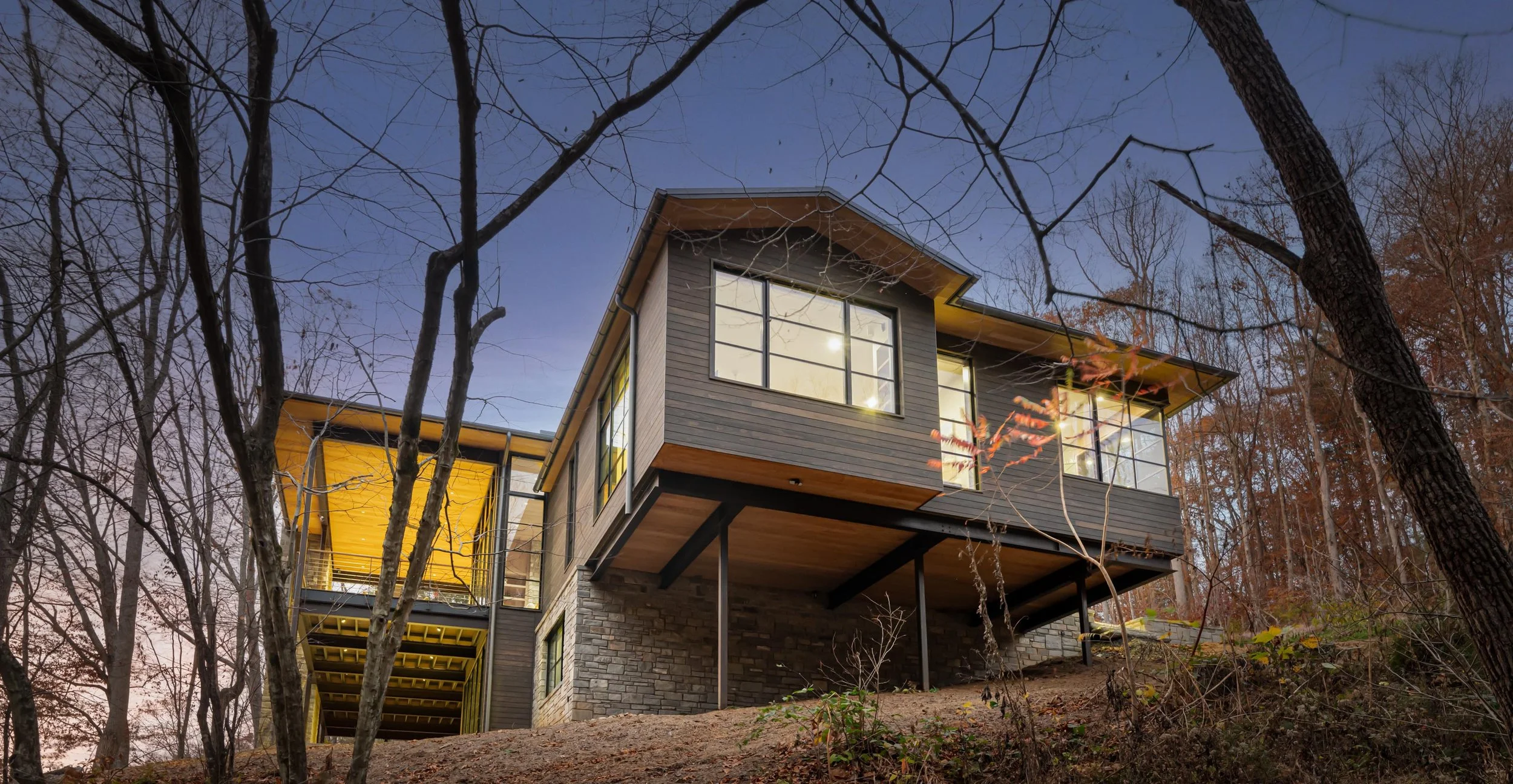 Modern house on a hillside surrounded by leafless trees during dusk with large windows and a yellow accent on the exterior