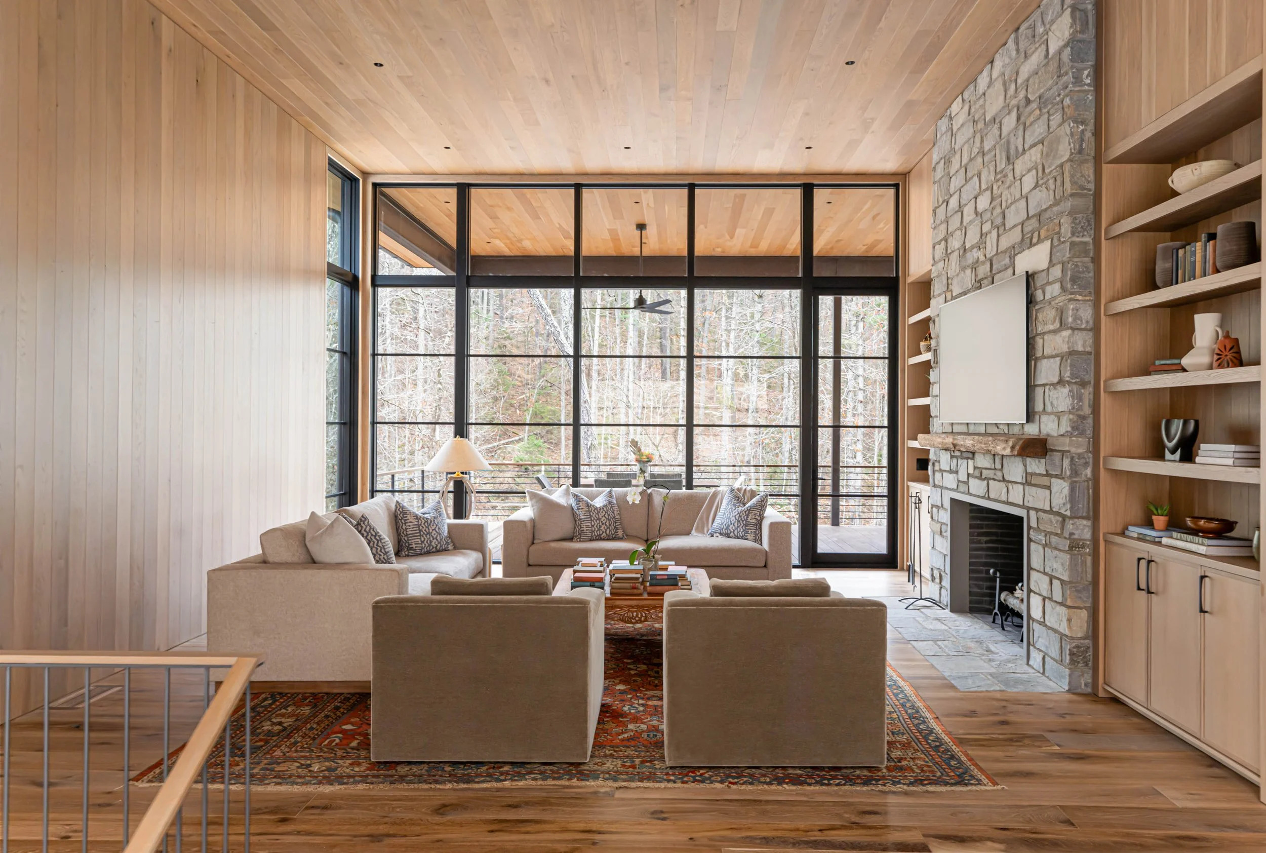 Bright living room with beige sofas and armchairs, wooden floors and ceiling, large glass windows overlooking a wooded outdoor area, a stone fireplace with a mounted TV, and built-in wooden shelves with decor items.