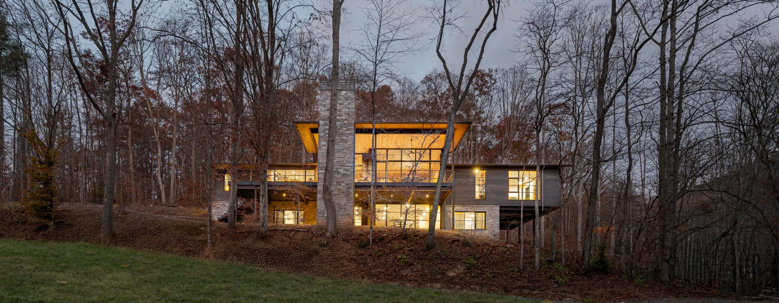 Modern house illuminated at dusk, situated on a wooded hillside with leafless trees surrounding it.