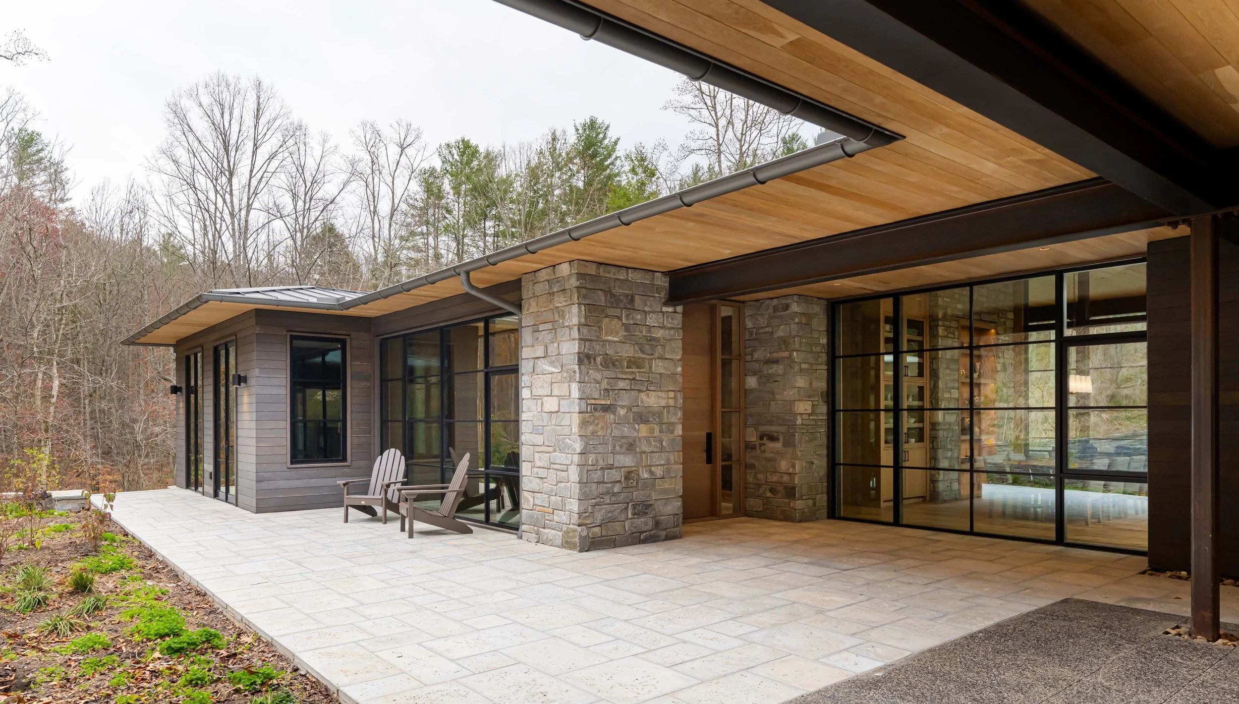 Modern house with stone and wood exterior, large glass windows and a porch with wooden chairs, surrounded by trees.