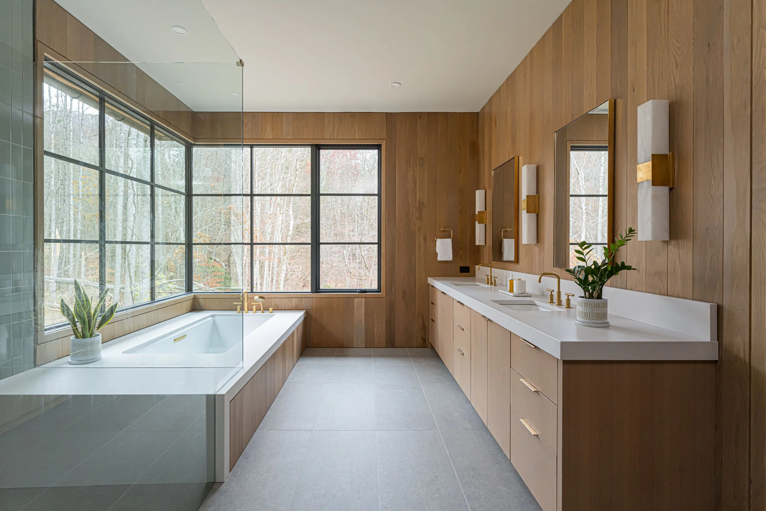 Modern bathroom with wooden walls, large window overlooking trees, white bathtub with gold fixtures, double vanity with matching gold fixtures, mirrors, wall sconces, potted plant, and neutral floor tiles.