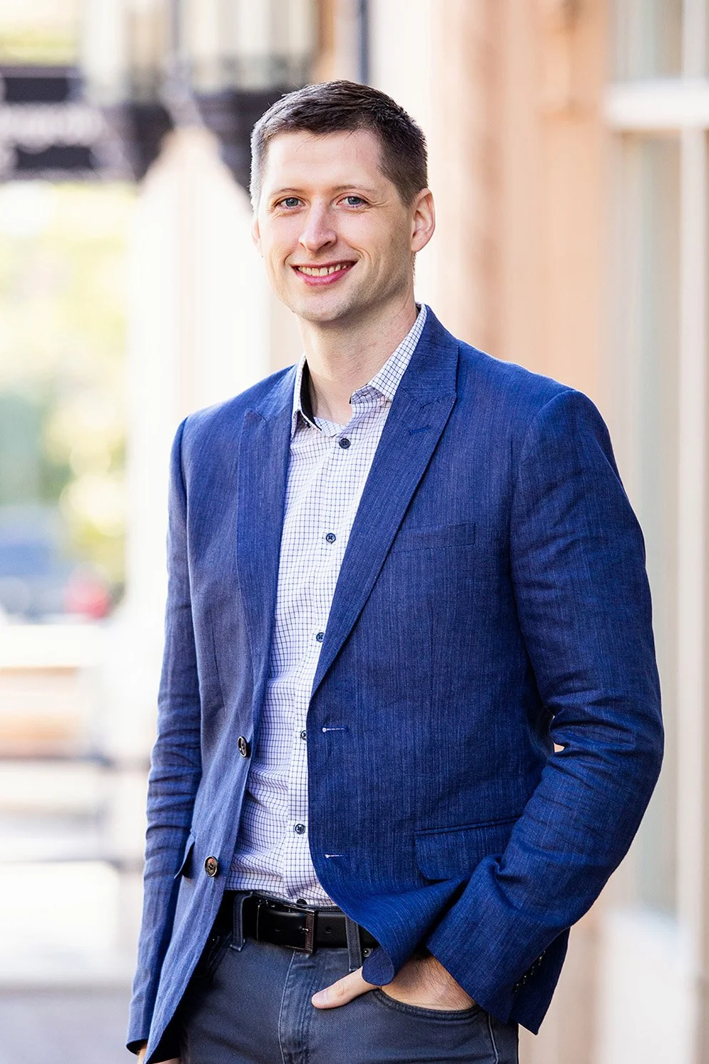 A man wearing a blue blazer and a checkered shirt standing outdoors, smiling at the camera.