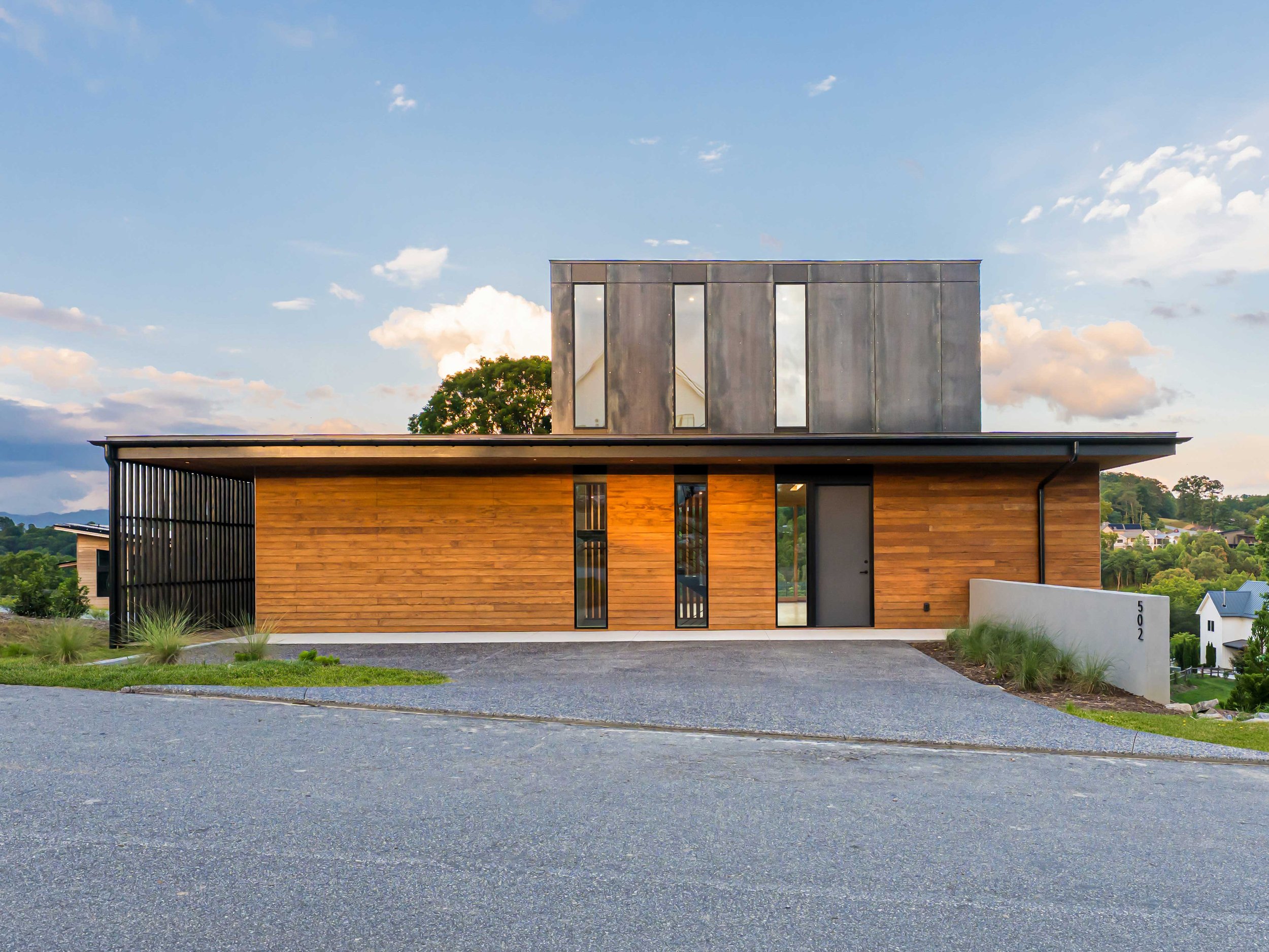 Modern house with wooden and metal exterior, gravel driveway, situated on a hill with a view of greenery and neighboring houses, under a partly cloudy sky.