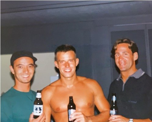 Three men standing indoors, smiling, and holding beer bottles. The shirtless man is in the center, with the other two men on each side.