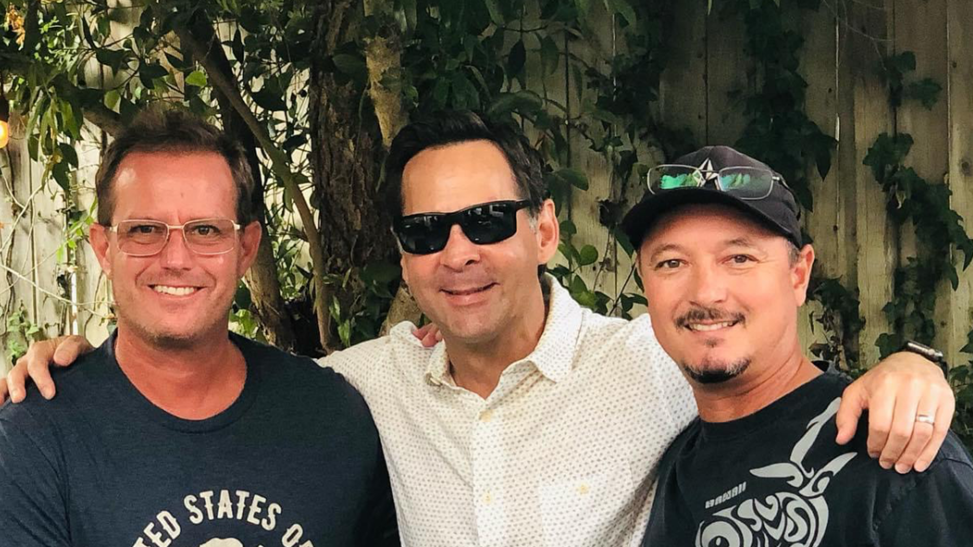 Three men smiling outdoors, standing close together with arms around each other, with trees and a wooden fence in the background.