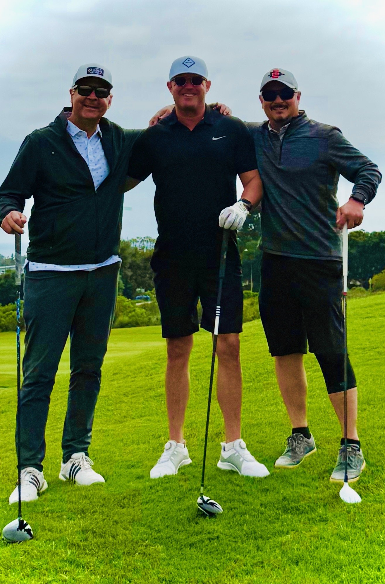 Three men standing on a golf course, posing with golf clubs, smiling, dressed in casual golf attire, with green grass and trees in the background.