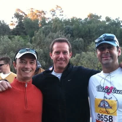 Three men smiling outdoors, with trees in the background. The man on the left is wearing a red jacket and a white cap. The man in the middle is wearing a black shirt. The man on the right is wearing a white T-shirt with a logo, sunglasses, and a cap.
