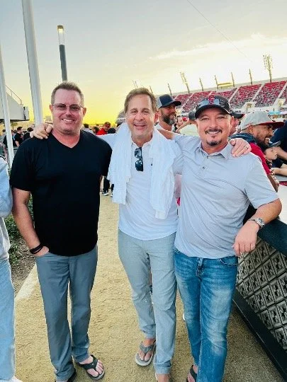 Three men standing together on a dirt surface at an outdoor event, smiling with arms around each other, with a crowd and stadium seats in the background.