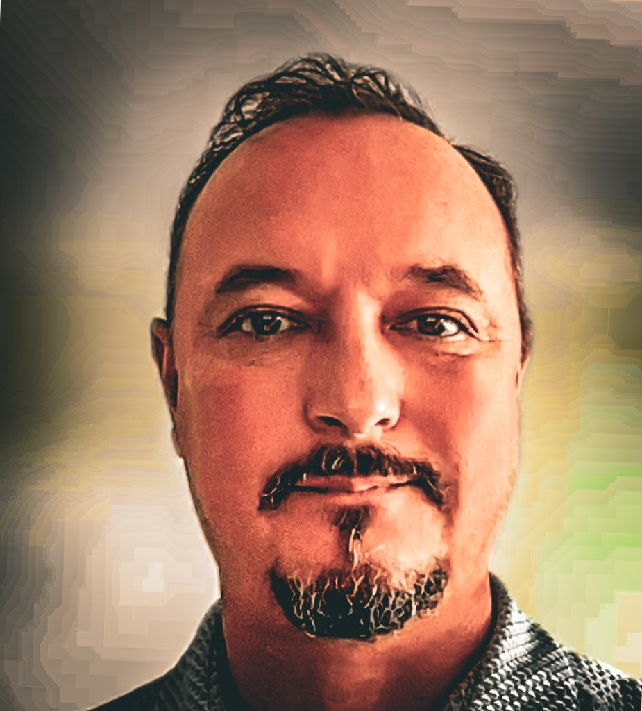 A close-up portrait of a middle-aged man with dark hair, a goatee, and a moustache, wearing a patterned shirt, smiling slightly against a blurred background.