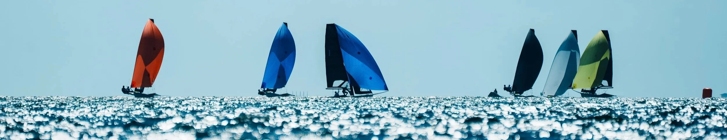 Several sailboats with colorful sails (orange, blue, black, white, yellow-green) sailing on the water with sunlight reflecting off the surface.