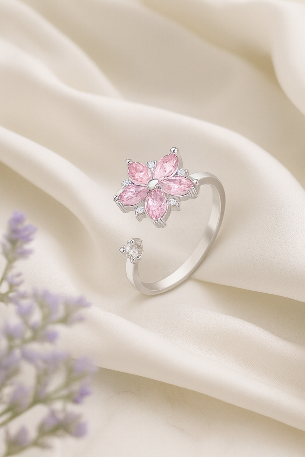 Silver ring with a pink flower-shaped cluster of pink gemstones and small clear gemstones, resting on cream-colored satin fabric, with purple lavender flowers in the foreground.