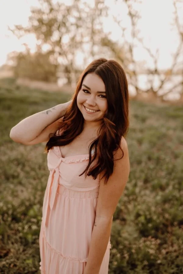 A woman with short blonde hair, wearing a white short-sleeved button-up blouse tied at the waist and tan pants, standing outdoors in front of trees and greenery, smiling at the camera.