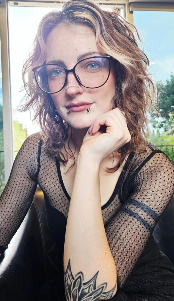 A woman with dark hair wearing a black sleeveless dress with a slit standing outdoors in front of a wire fence and plants.