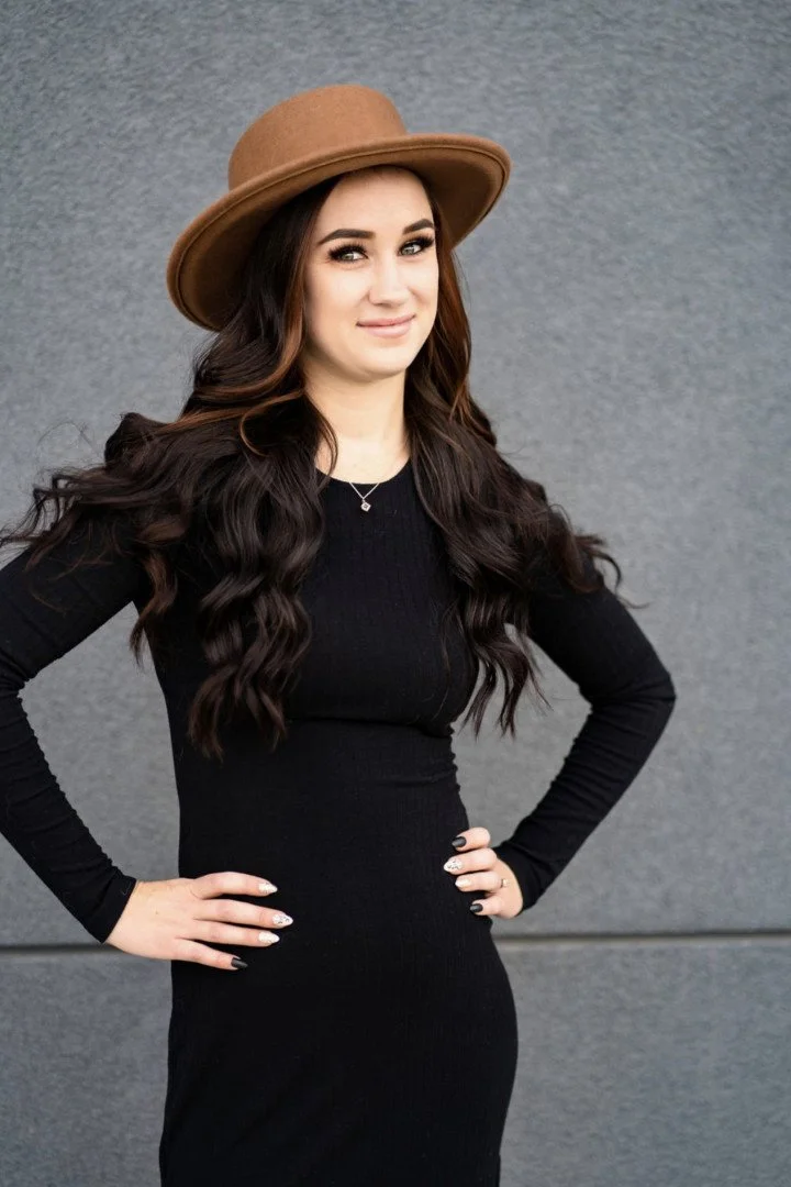 A woman with long, curly brown hair wearing a white sleeveless top and ripped jeans, sitting outdoors in front of a metal fence with some vines.