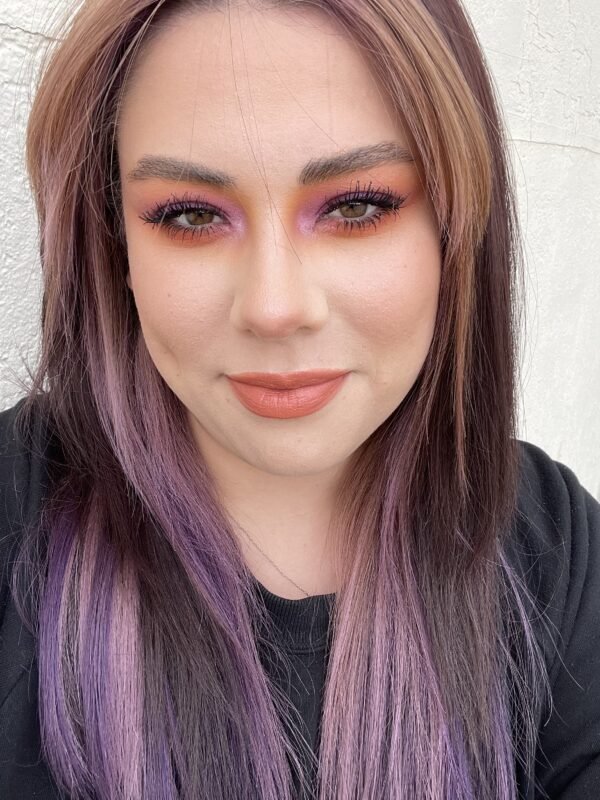 A woman with curly brown hair, wearing a black suit and red lipstick, standing outdoors in front of a rusty metal fence.
