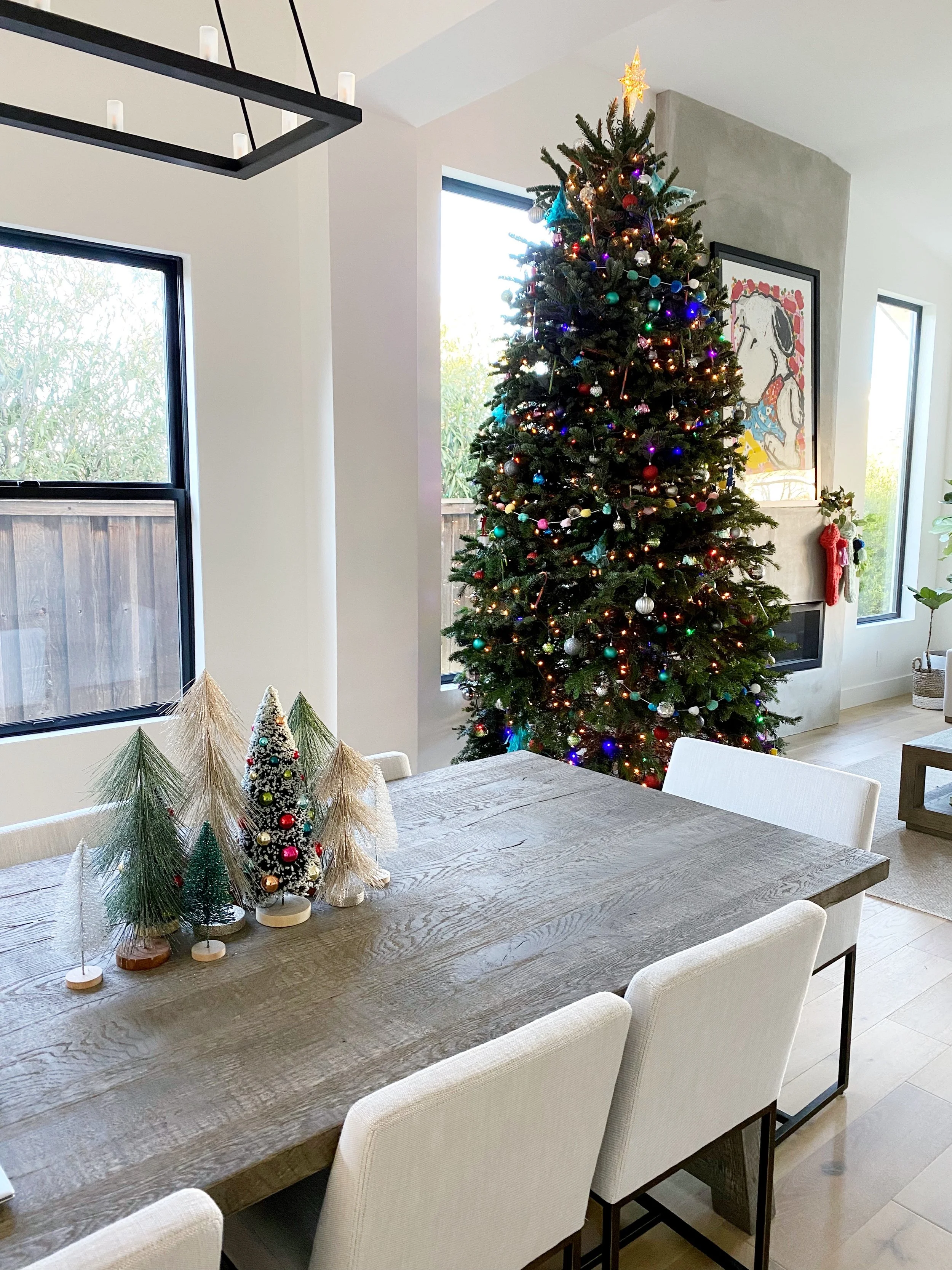 Living room with decorated Christmas tree, holiday decorations on fireplace, and dining table with miniature trees.