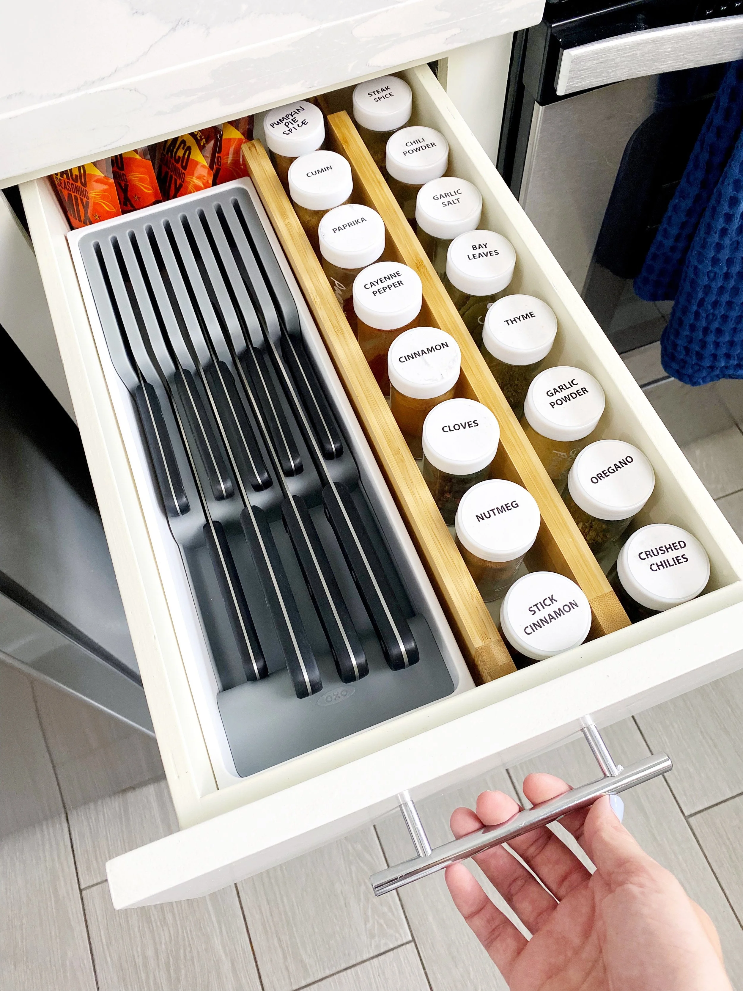 Open kitchen drawer with labeled spice jars, a bamboo spice rack, and black utensils on a gray tray.