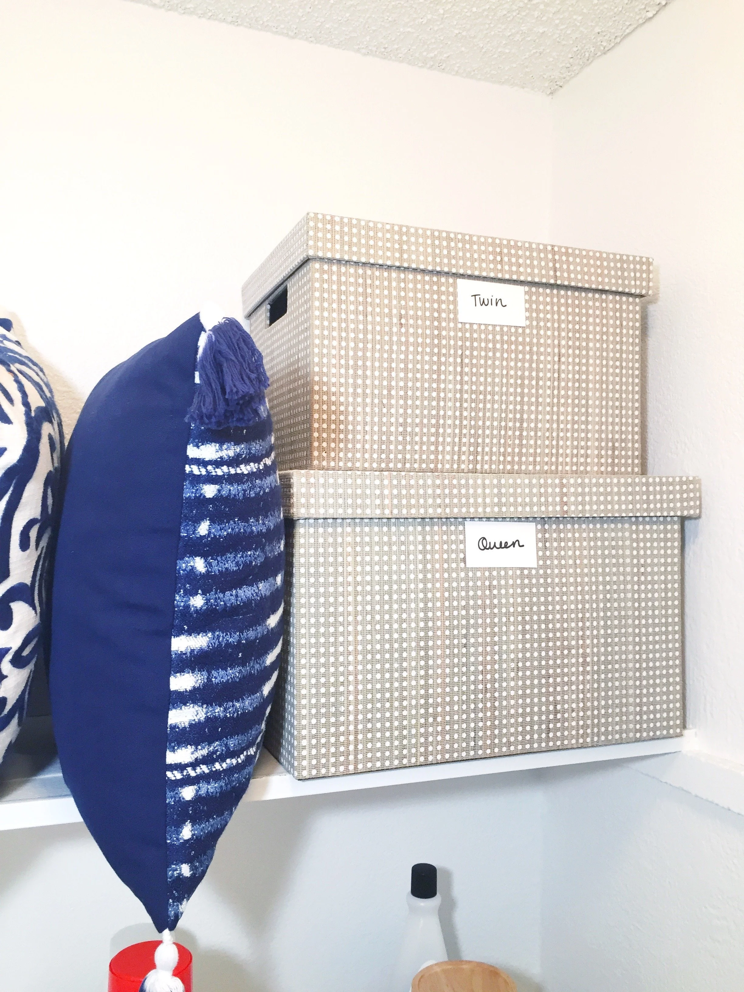 Two beige storage boxes with white polka dots on a white wall shelf, labeled "Twin" and "Queen", next to a blue patterned cushion and other household items.