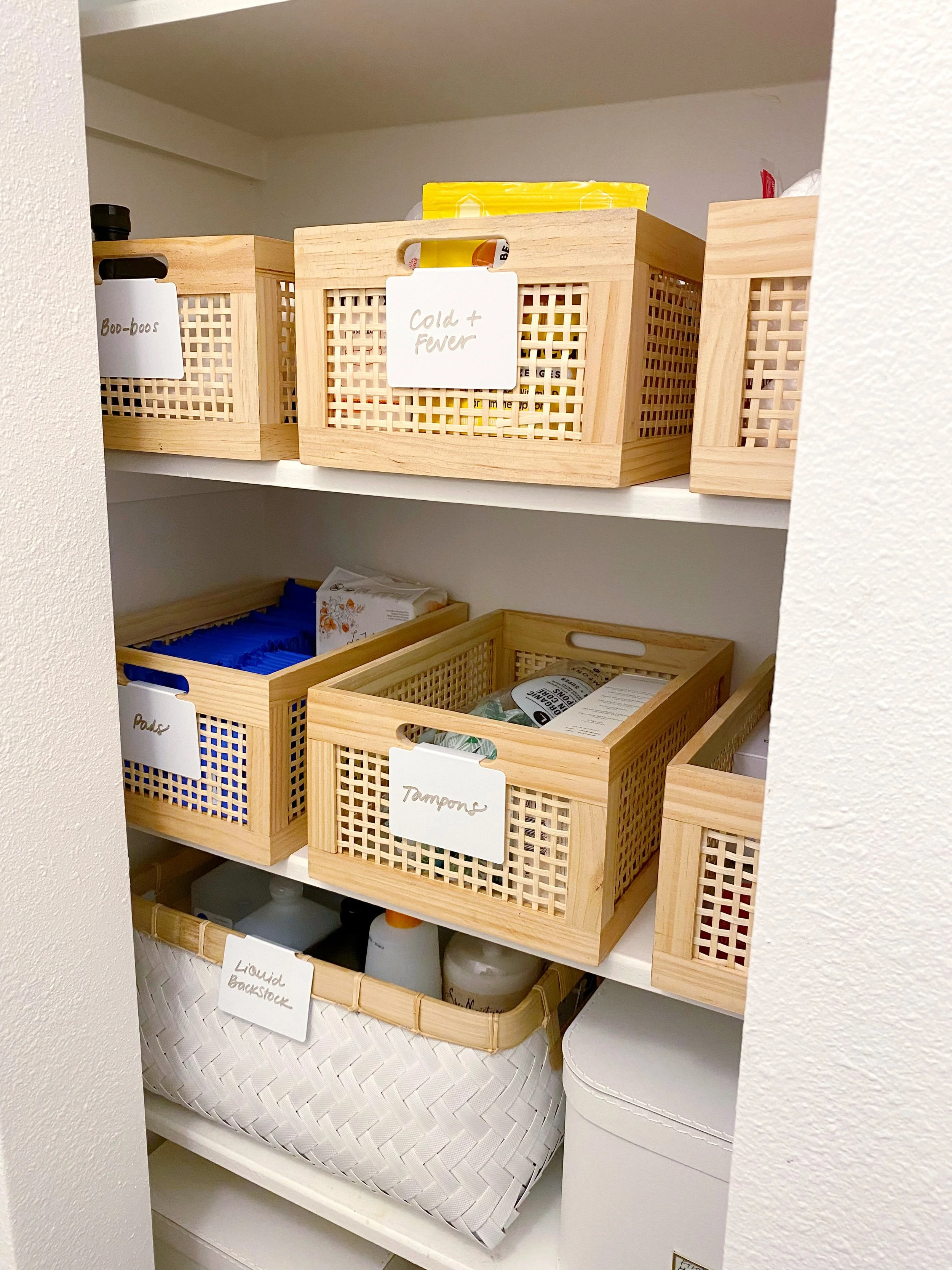 Closet shelf with organized storage baskets labeled 'Boo-boos,' 'Cold + Fewer,' 'Pads,' 'Tampons,' and 'Liquid Backstock' containing various health and hygiene supplies.