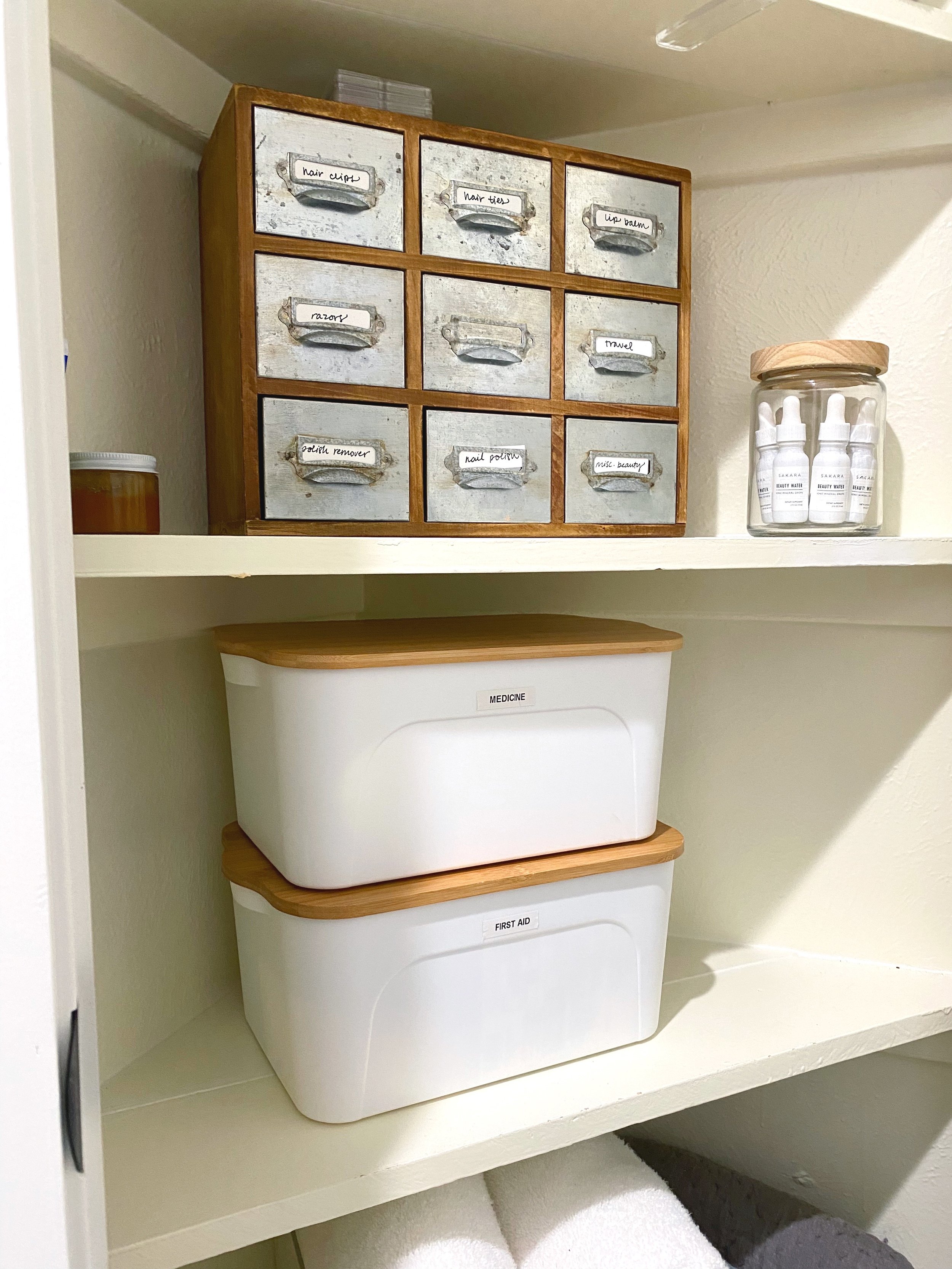 A wooden cabinet with labeled small drawers for organizing beauty products, a jar with skincare bottles, and two labeled plastic storage boxes for medicine and first aid in a closet or bathroom shelf.
