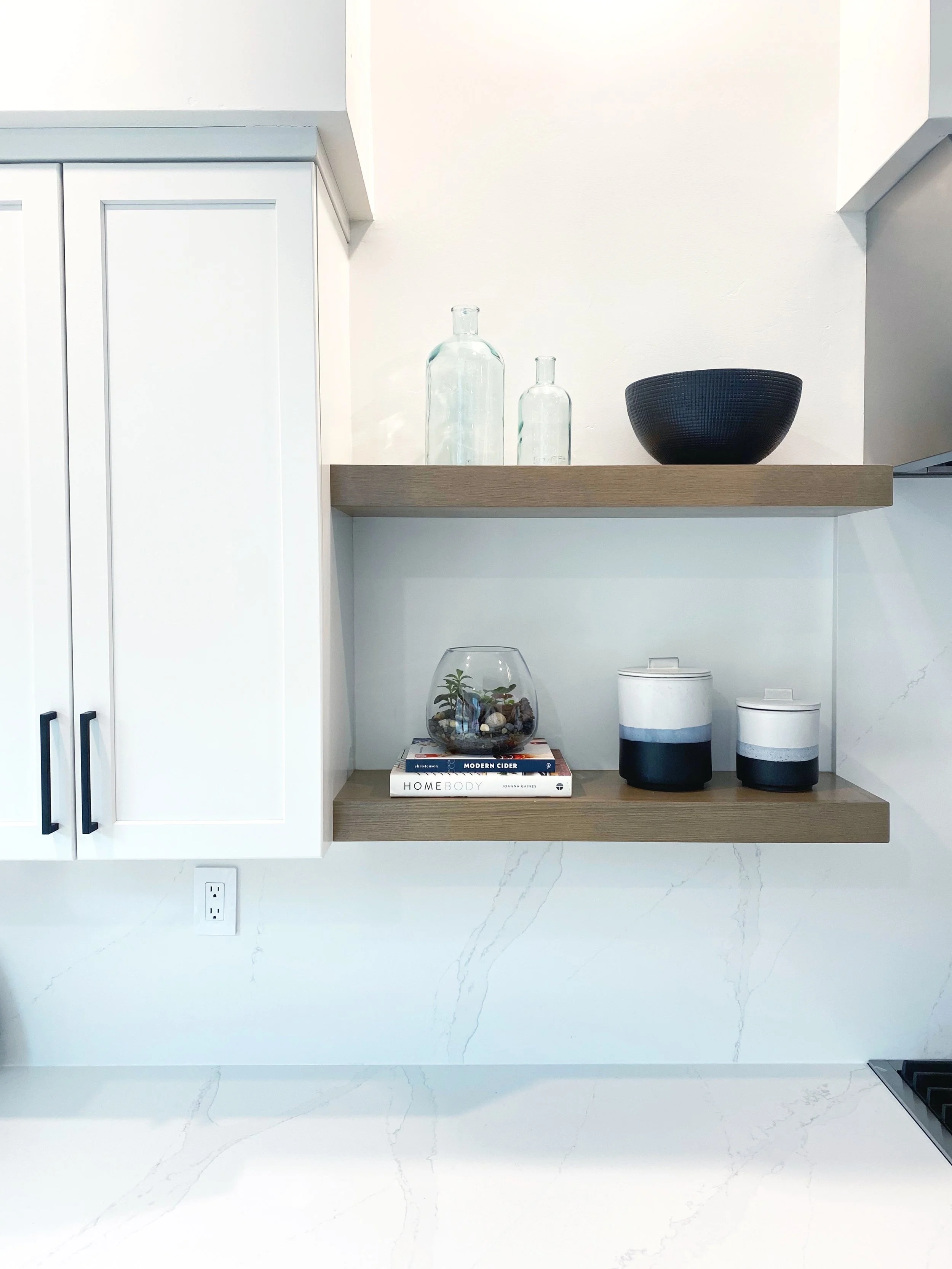 Open kitchen wall shelves with glass bottles, a black bowl, a small terrarium, and containers, with a white cabinet on the left and marble countertops.