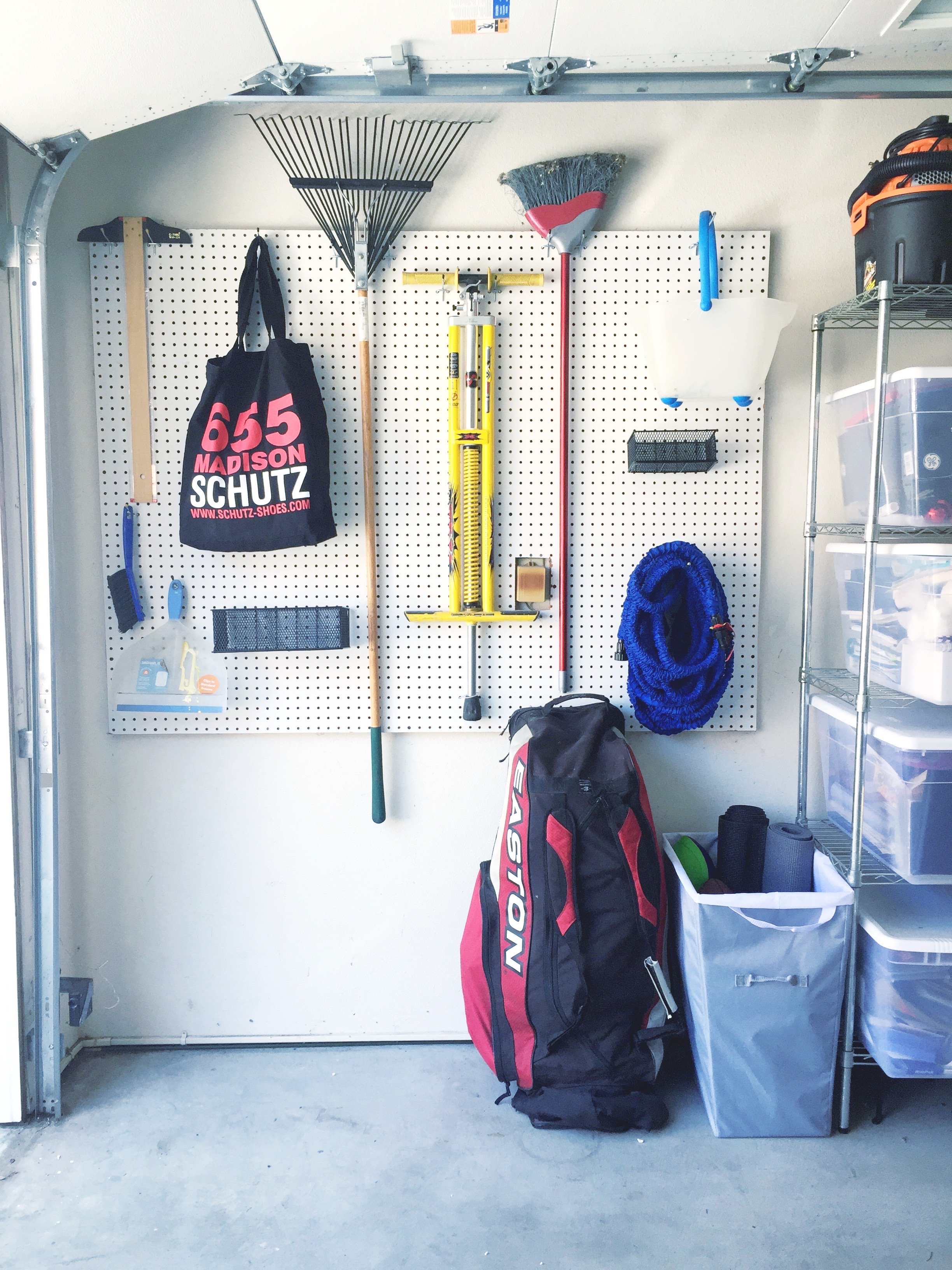 Garage door with tool pegboard holding a broom, rake, mop, and hanging bag, with a golf bag and storage bins on the floor.
