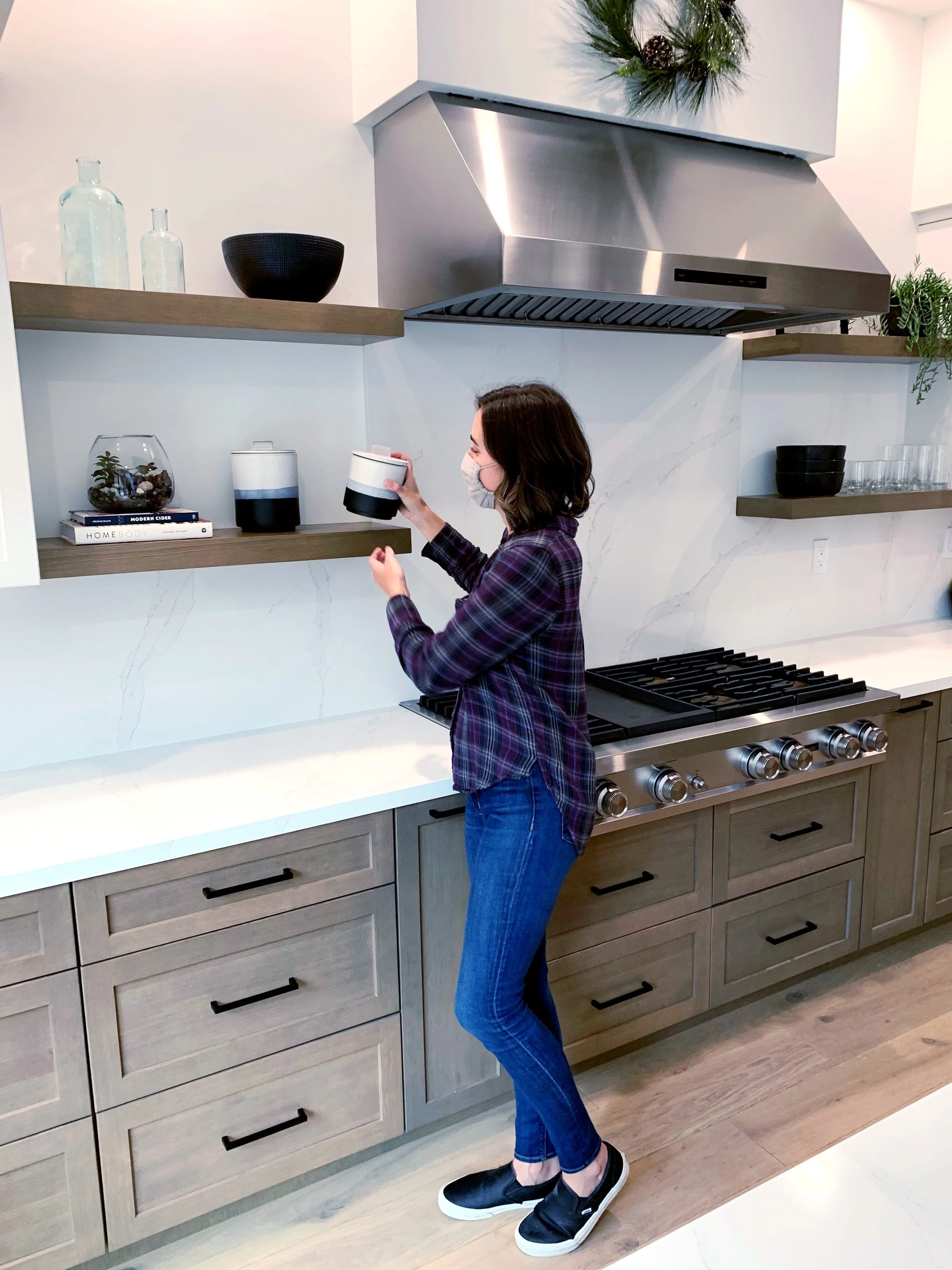 A woman in a face mask wearing a plaid shirt and jeans is standing in a modern kitchen, holding a white and black container, next to a stove with a stainless steel range hood and open shelves with decorative items and bottles.