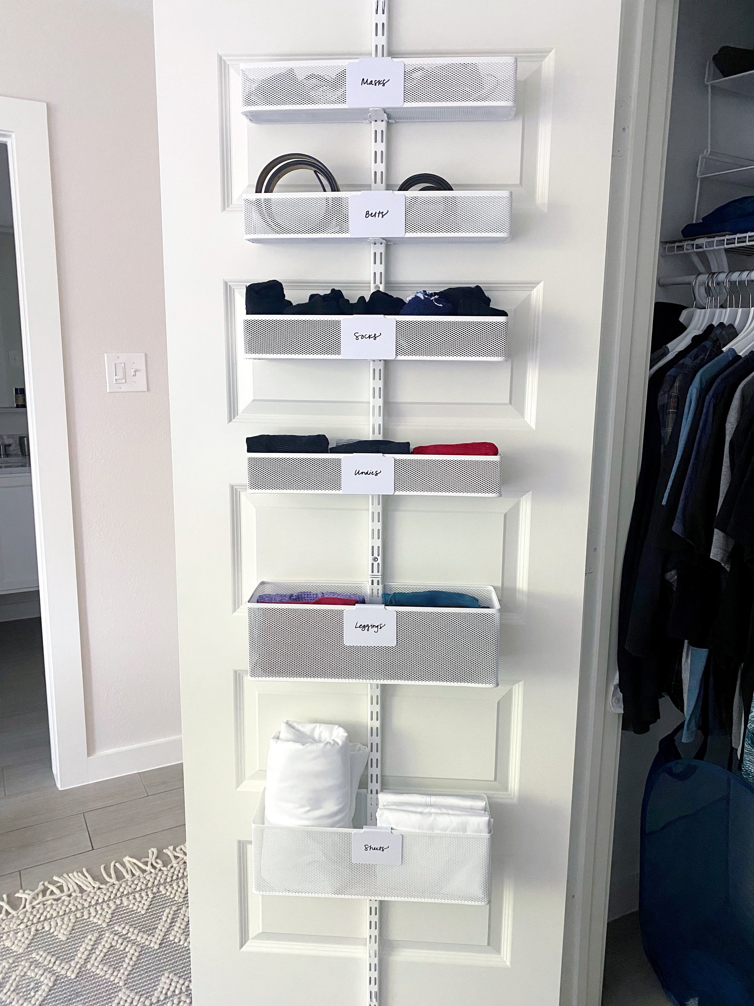 Vertical storage organizer with labeled white mesh baskets holding various clothing items such as underwear, socks, and shirts, mounted on a white door.