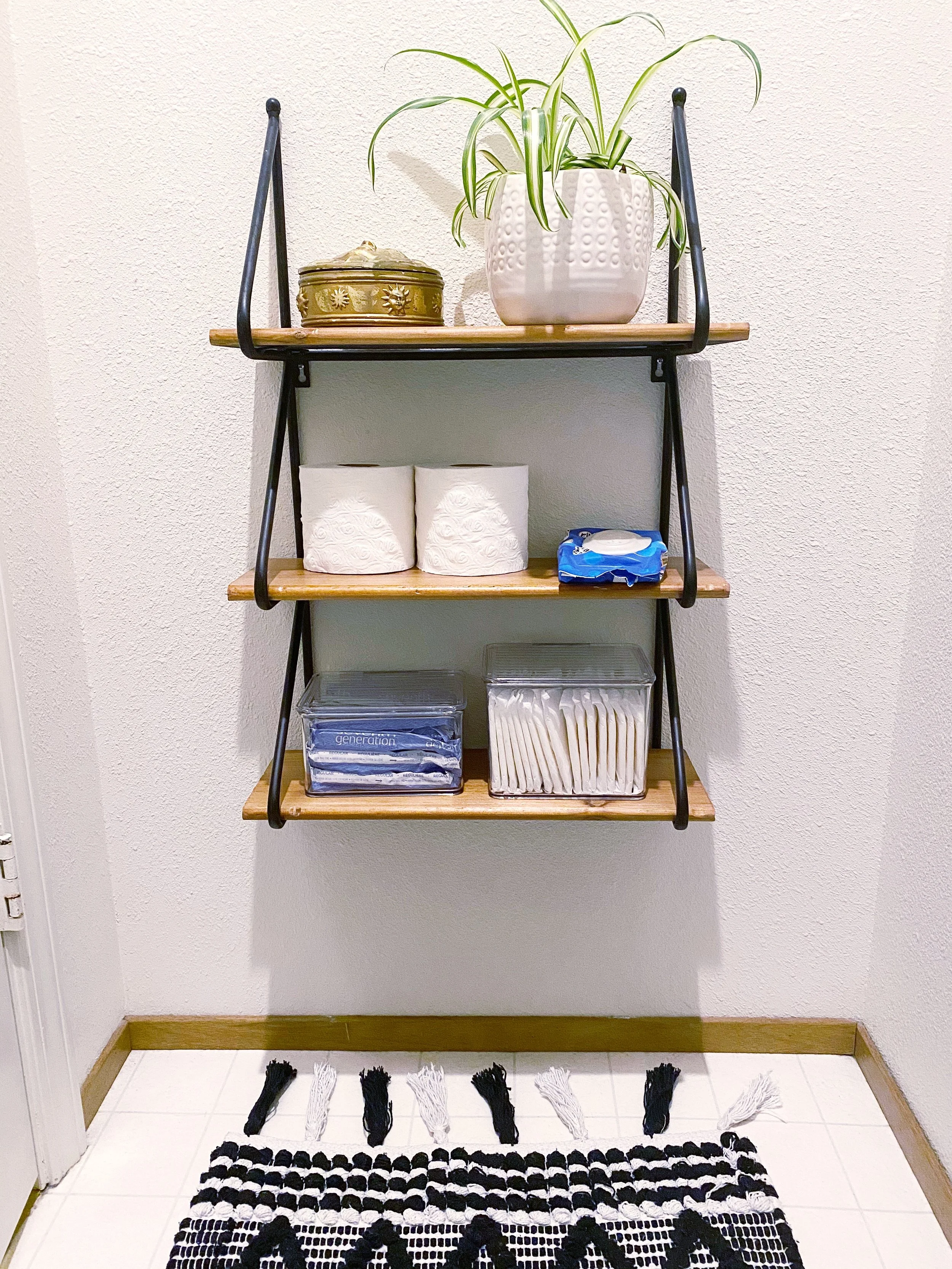 Wall-mounted wooden and metal shelves with decorative items including a potted plant and tissue boxes. Below a black and white woven rug with tassels is on a tiled floor.