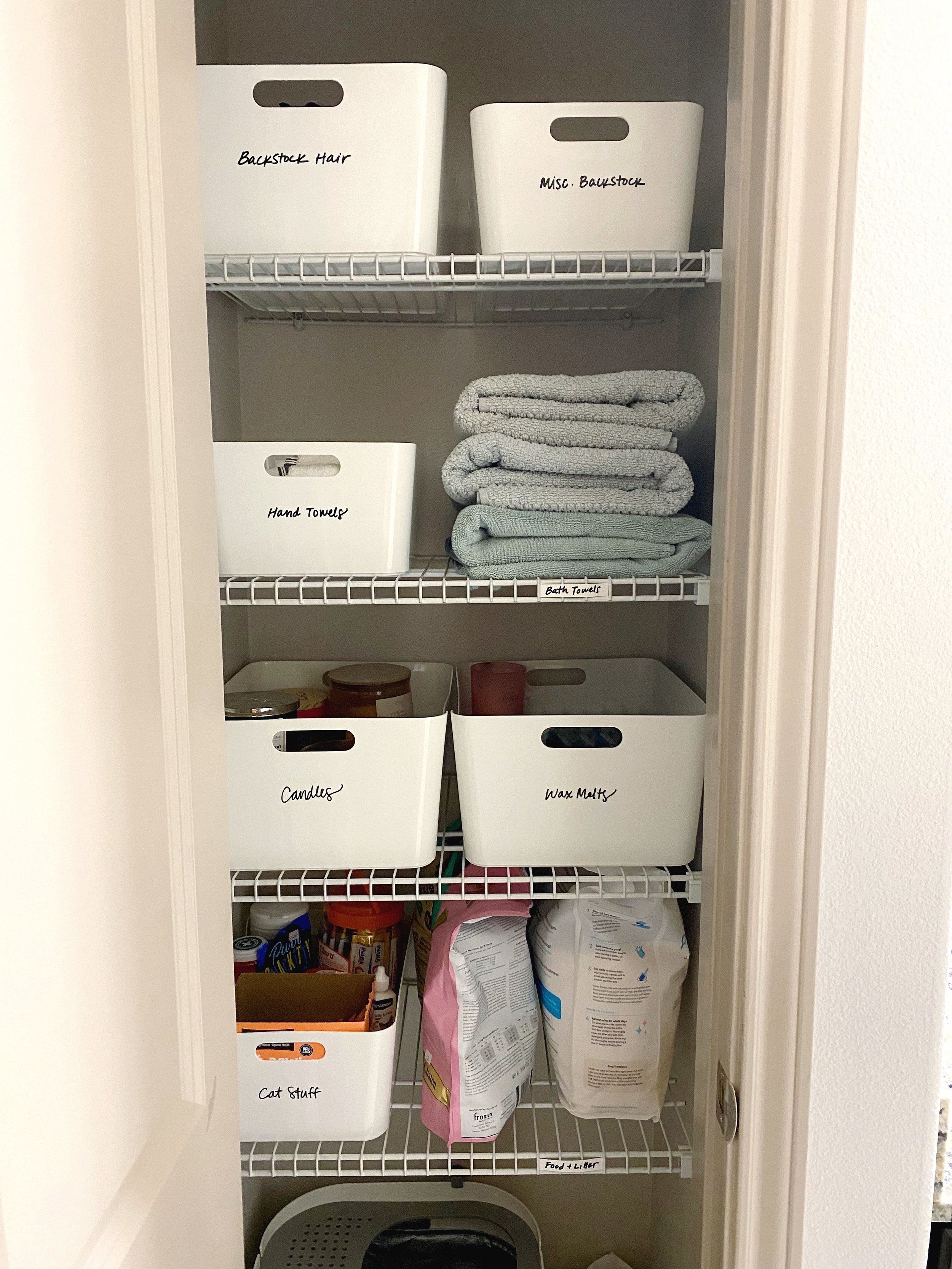 Inside a closet with shelves holding white labeled storage bins, towels, candles, wax melts, and cat supplies.