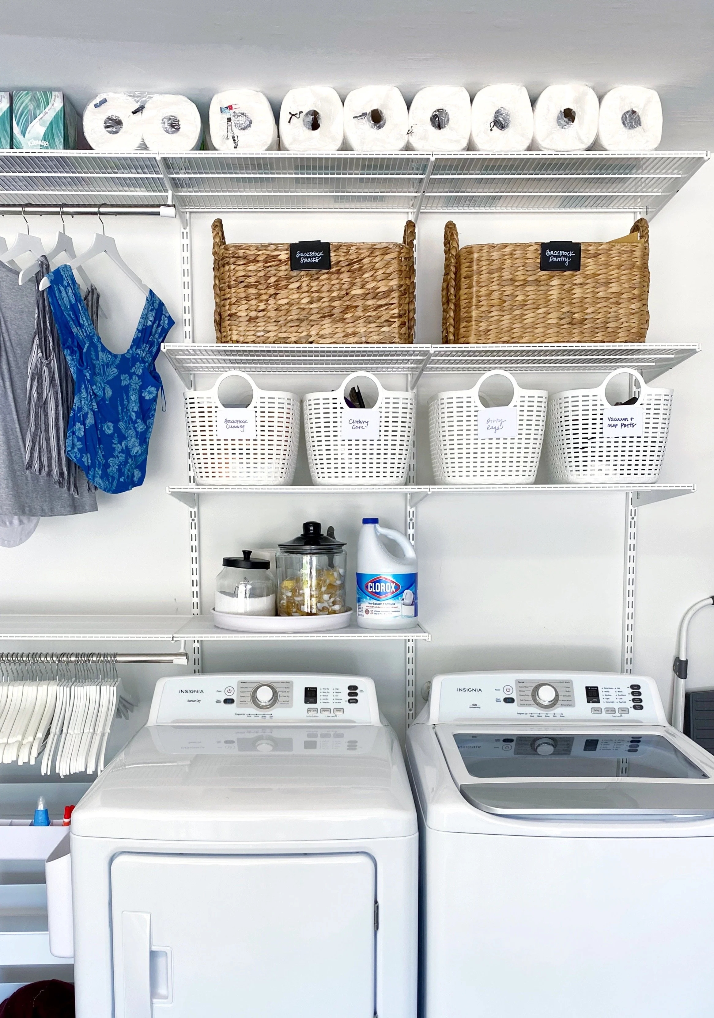Laundry room with two washing machines, storage shelves with laundry supplies, wicker baskets, white laundry baskets labeled for different purposes, bottles of cleaning products, and clothing hanging on racks.