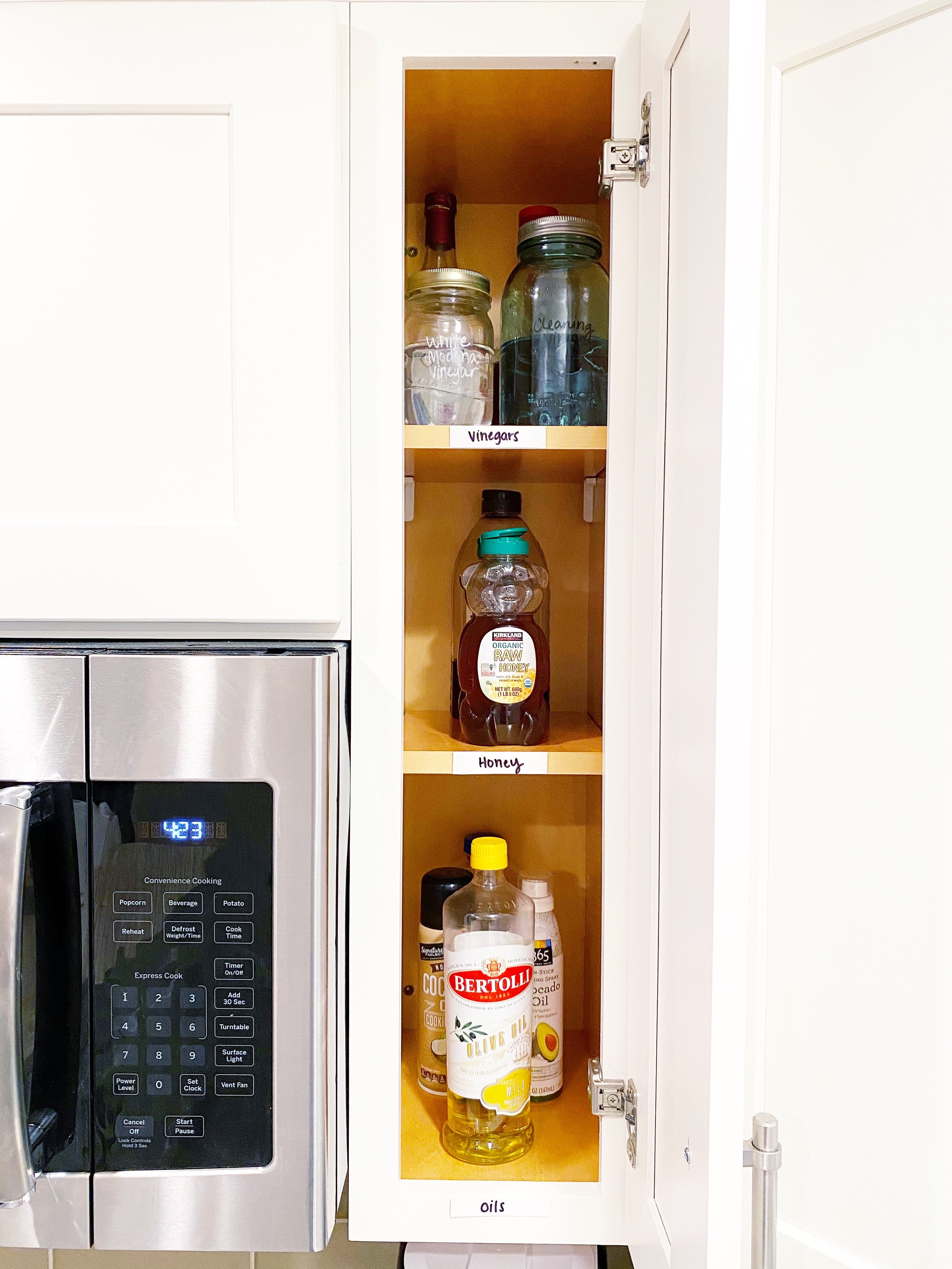 Open kitchen cabinet with three shelves containing jars labeled vinegar and honey, and bottles labeled oils, with a microwave oven to the left.
