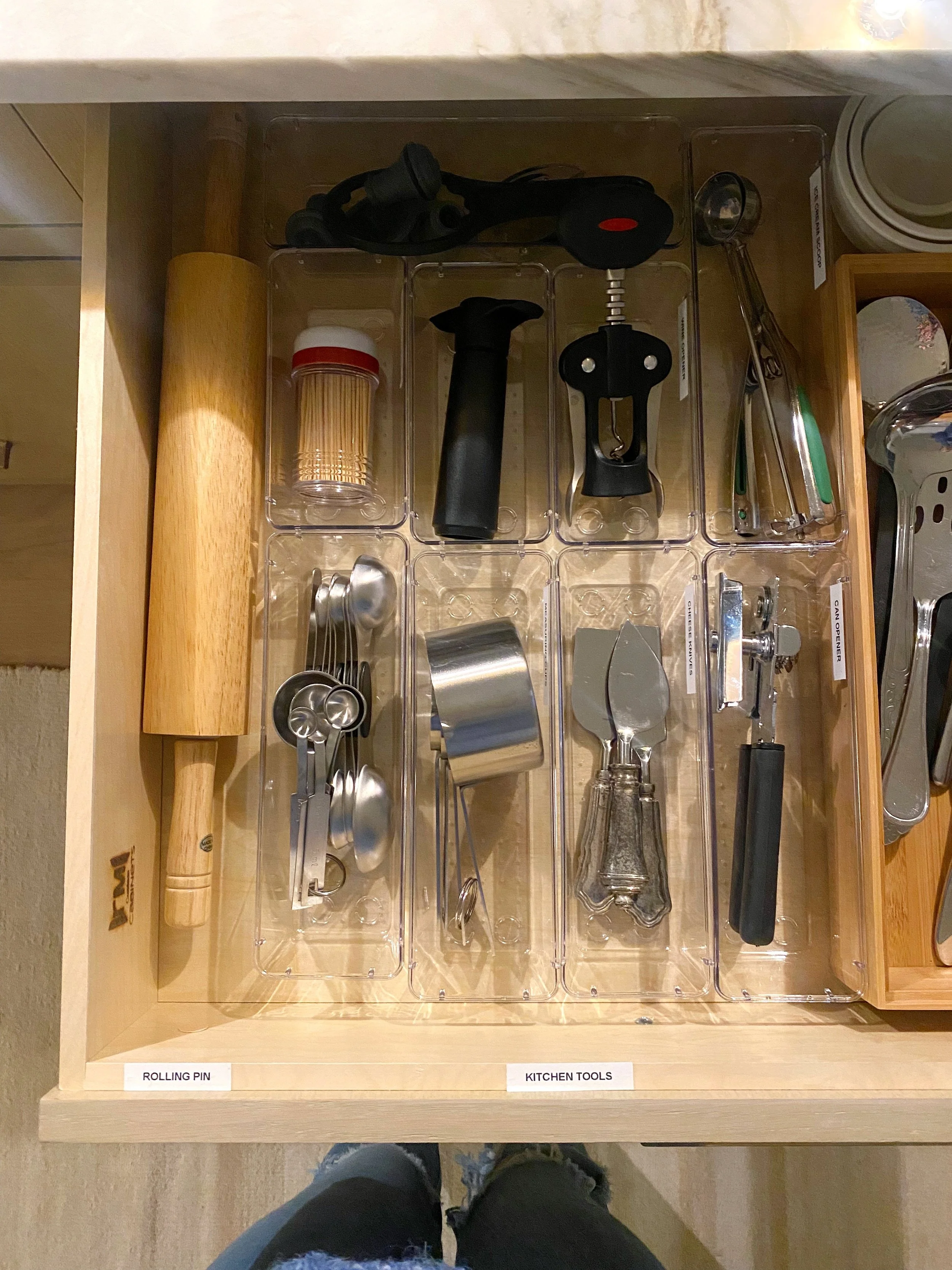Organized kitchen drawer with a rolling pin, kitchen tools, measuring spoons, a cup, and a handheld can opener.