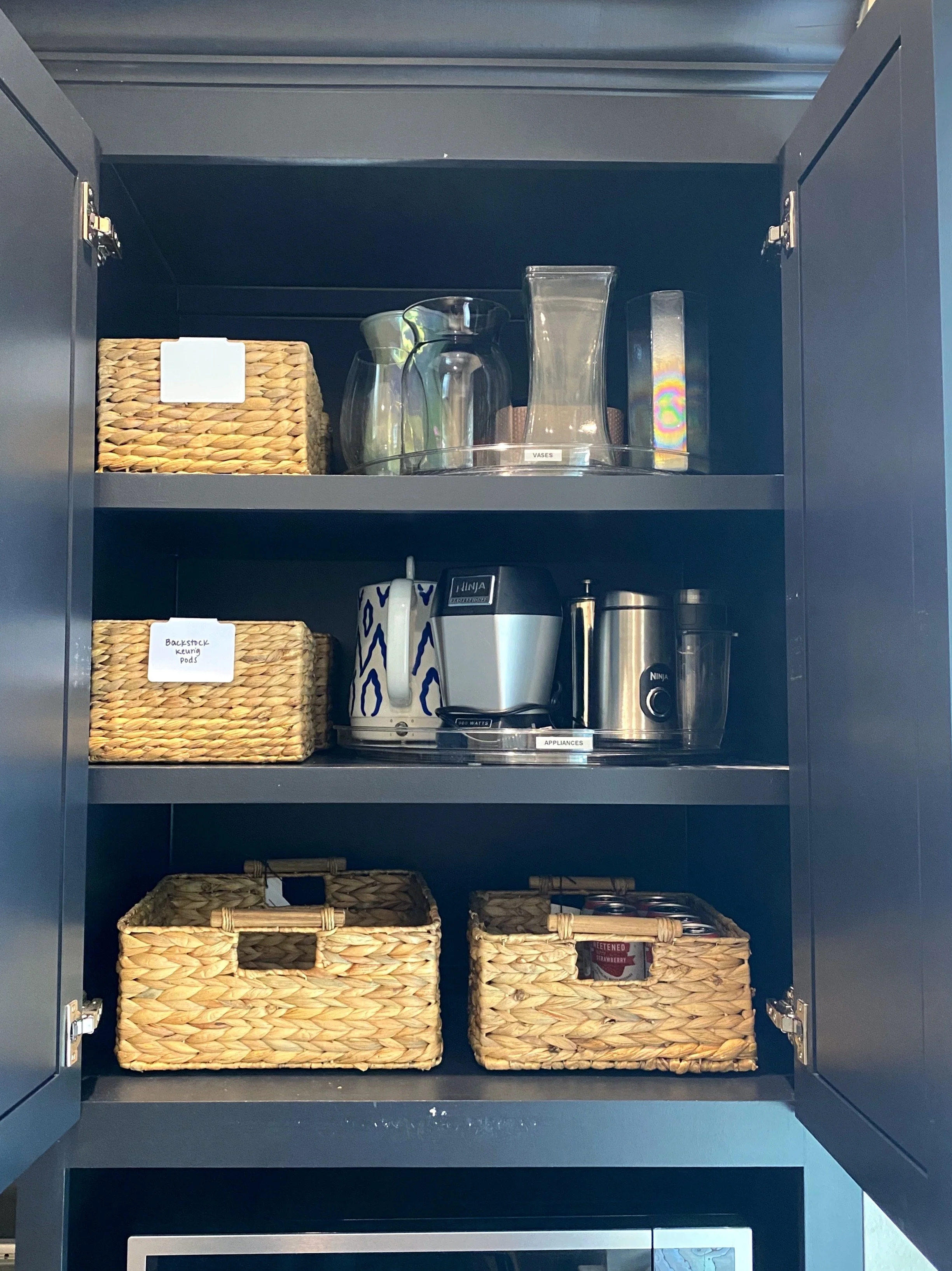 Kitchen cabinet with glassware, a coffee maker, a kettle, a blender, and woven baskets on shelves.