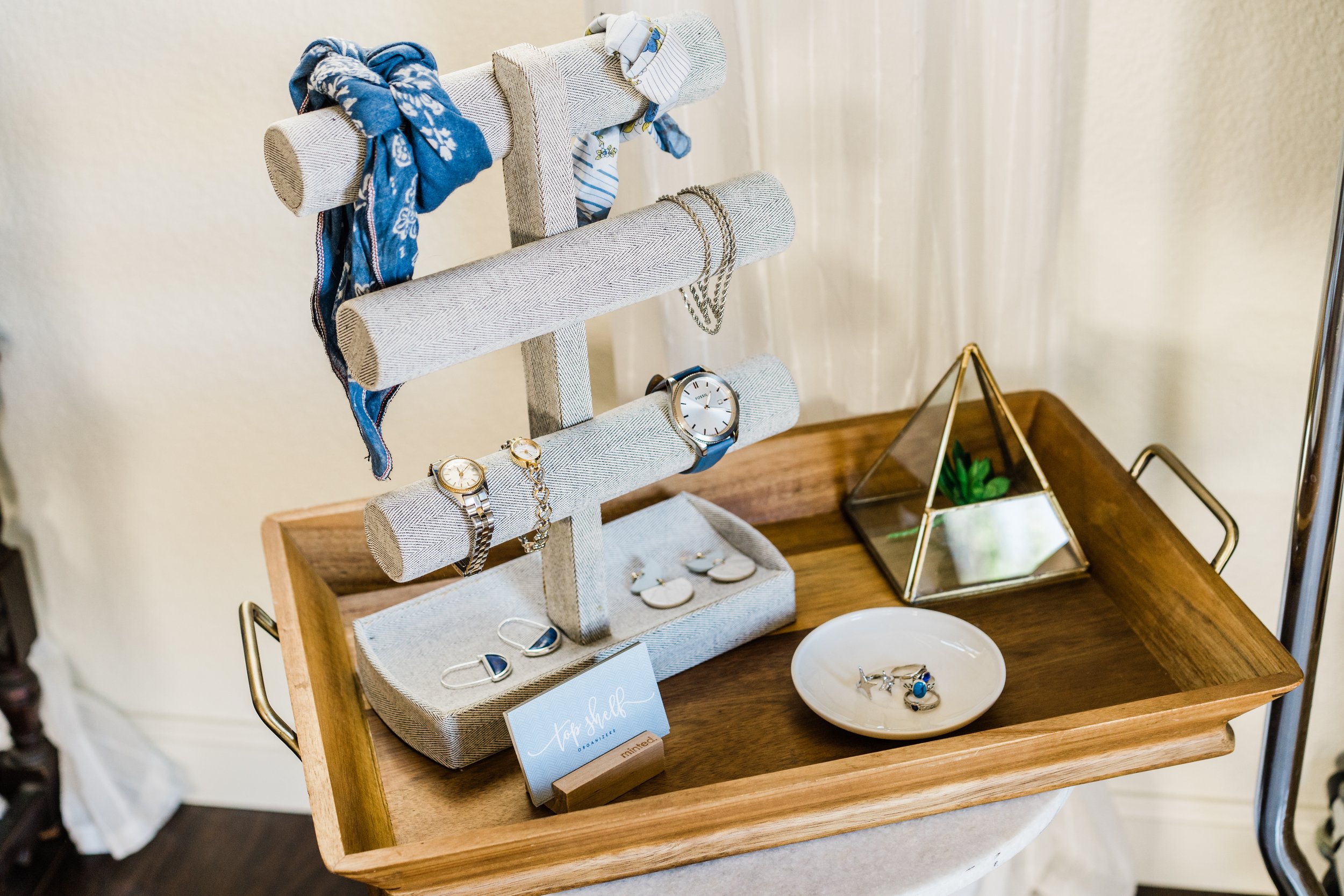 Jewelry display stand with watches, earrings, and necklaces on a wooden table, with a geometric glass terrarium and a white bowl holding rings nearby.