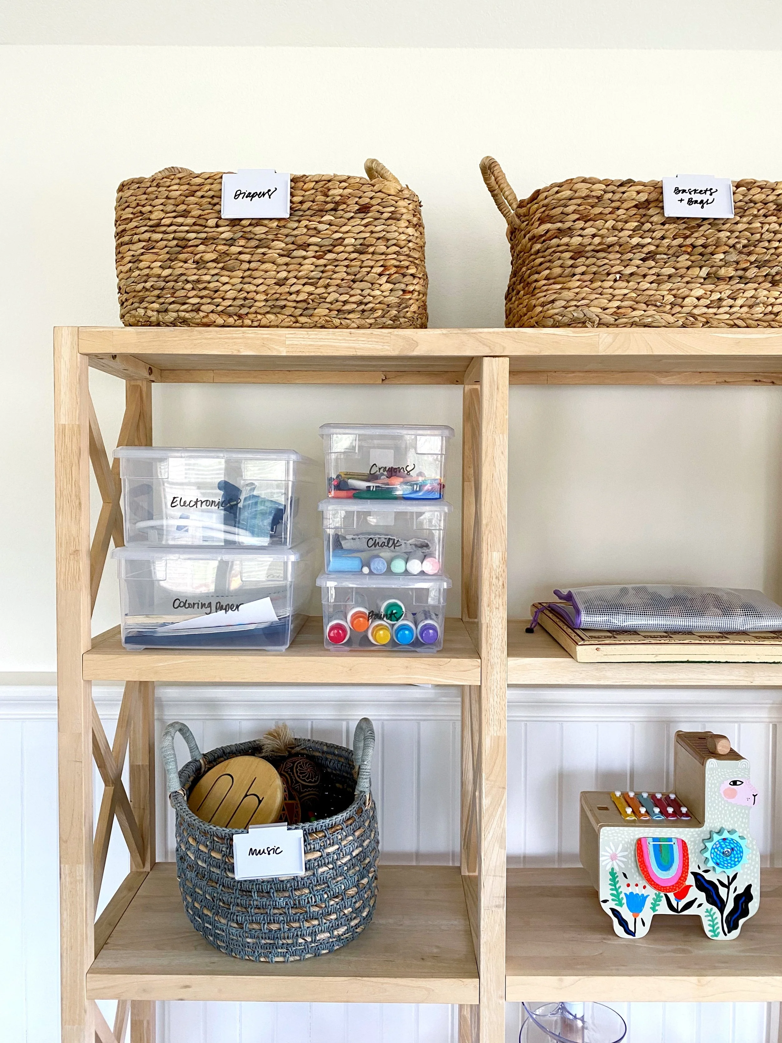 A wooden shelving unit with labeled storage bins and baskets containing children's toys, art supplies, and musical instruments. The top shelves have woven baskets labeled 'Diapers' and 'Baskets + Bows.' The middle shelves contain clear plastic bins l