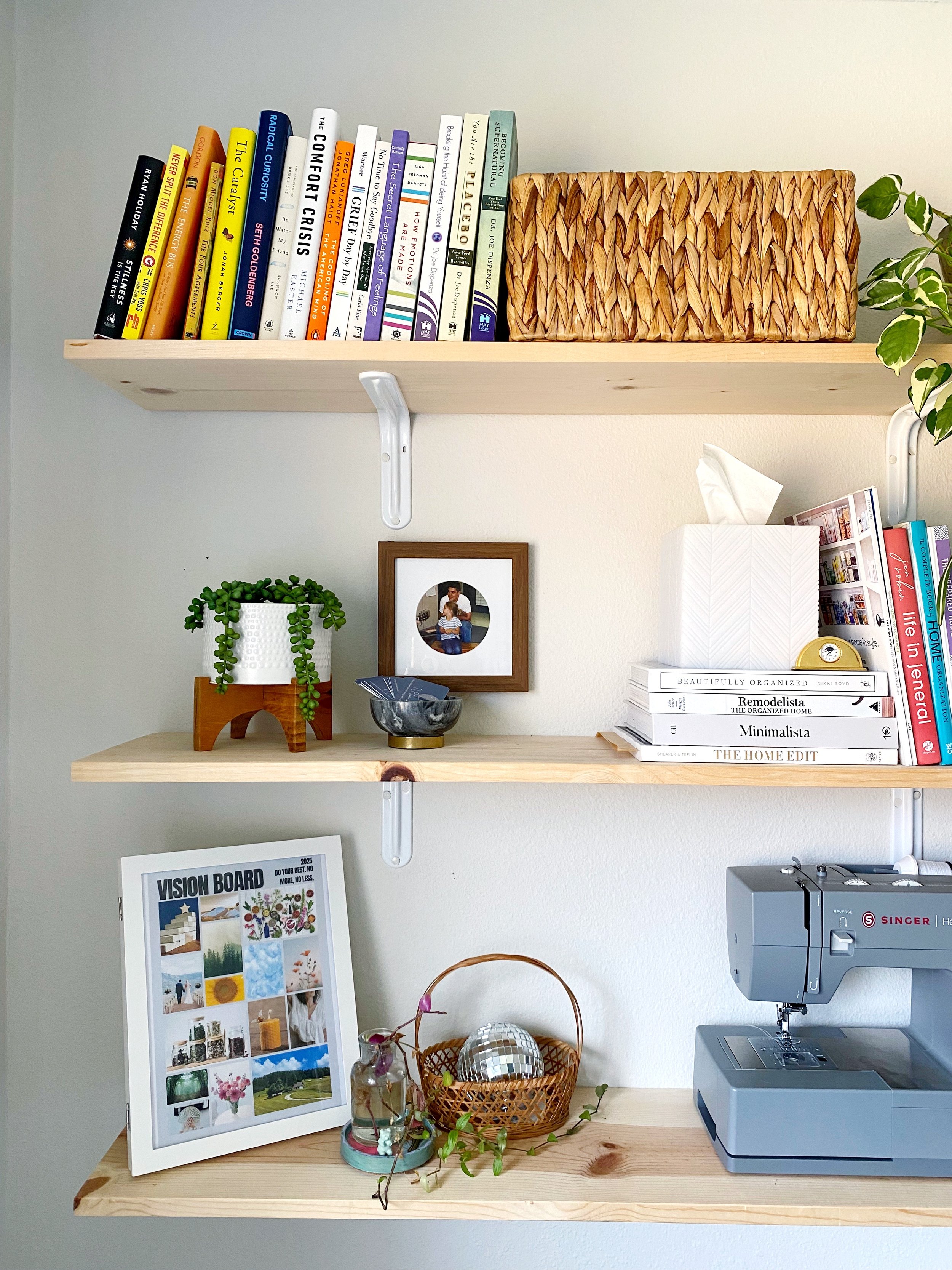 Two light-colored wooden shelves mounted on a white wall. The top shelf has books and a decorative woven basket. The middle shelf has a potted plant, a framed photo, a bowl, and stacked books. The bottom shelf has a framed collage, a small vase with 