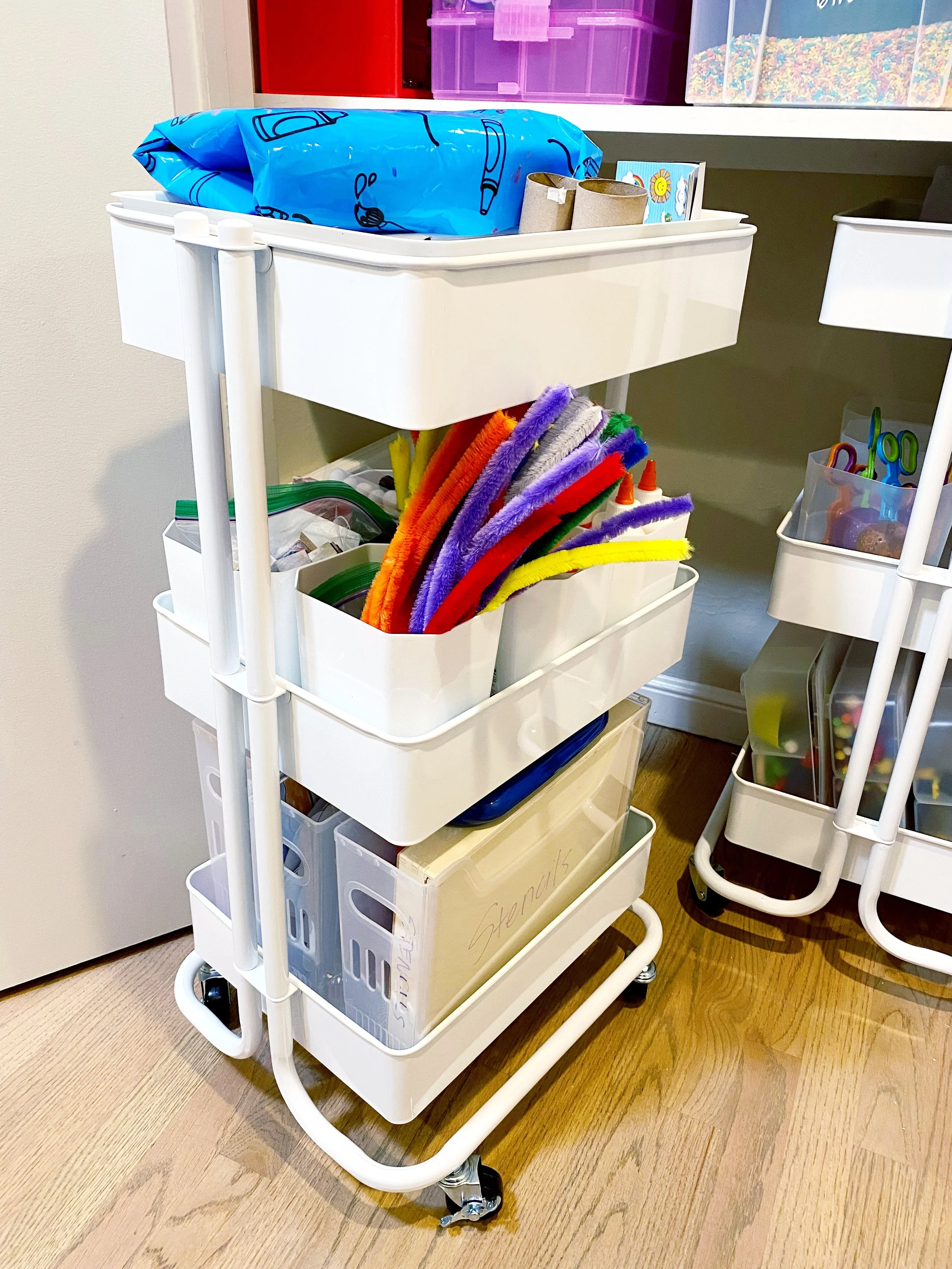 White rolling cart with multiple storage tiers containing craft supplies, including colorful pipe cleaners, scissors, and organizing containers, in a room with wooden floors.