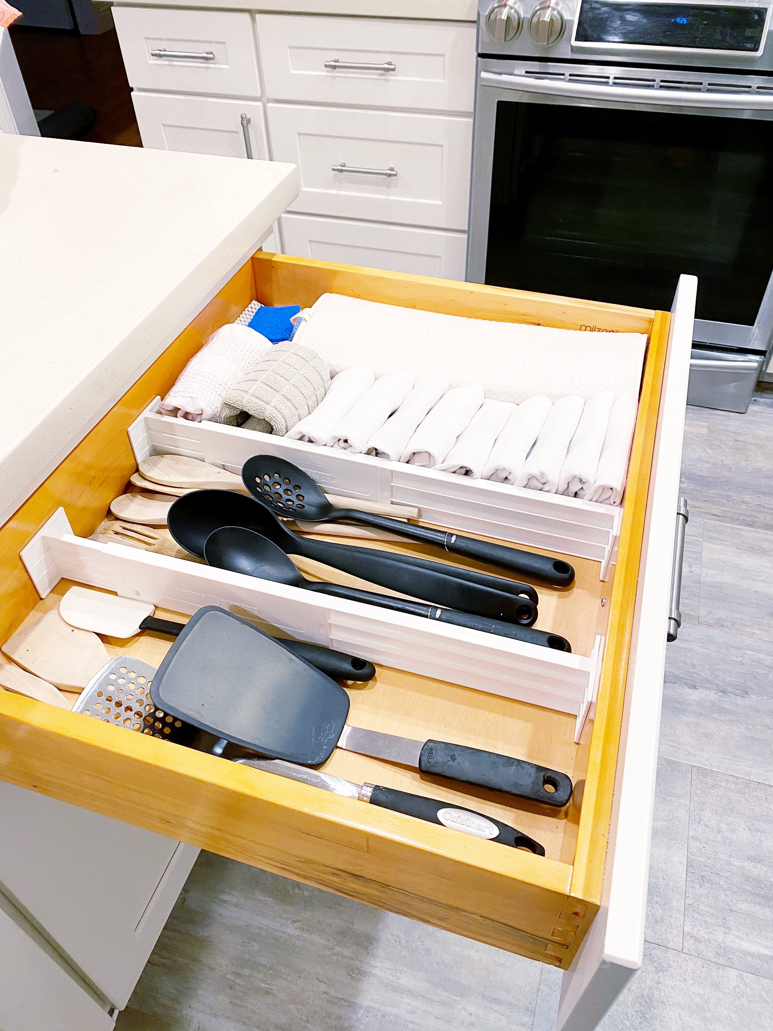 Kitchen drawer opened to reveal utensils after being organized, with a box of rolled-up cloths and cleaning cloths on top, surrounded by white cabinets, a gray stove, and a white countertop.