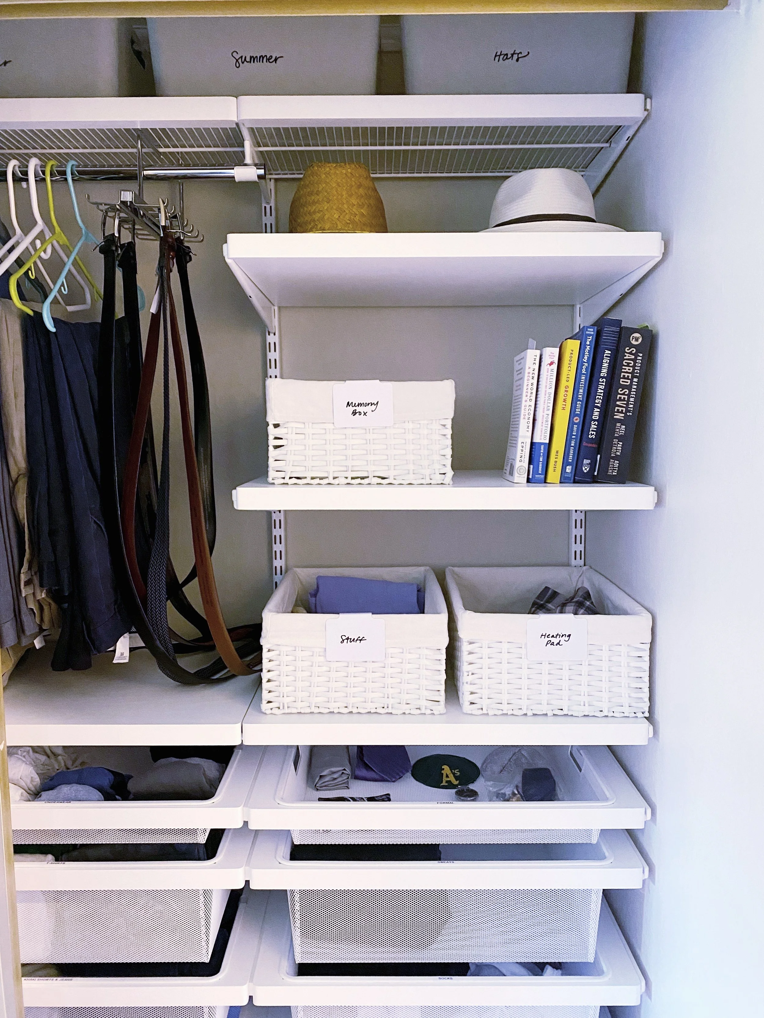 Closet with white shelves and baskets labeled for storage, containing hats, books, and clothing, with hanging belts and pants on the side.