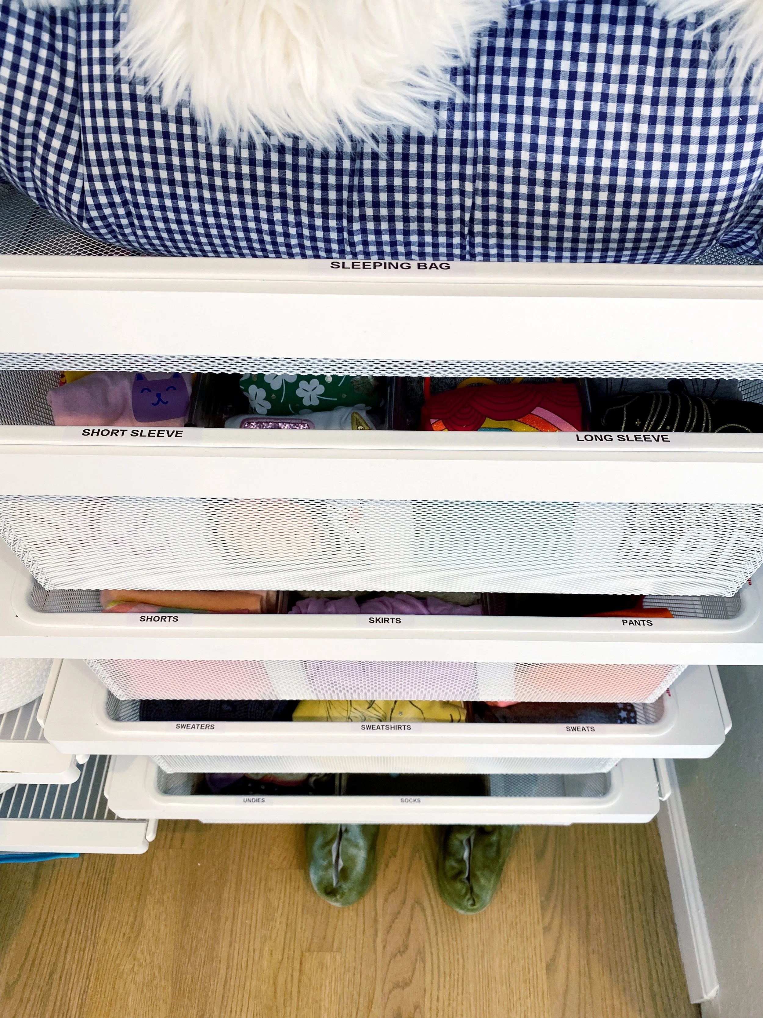 Inside a clothing storage unit with labeled sections, including sleeping bag, short sleeves, long sleeves, shorts, skirts, pants, sweaters, sweatshirts, undies, and socks, with green shoes visible at the bottom.