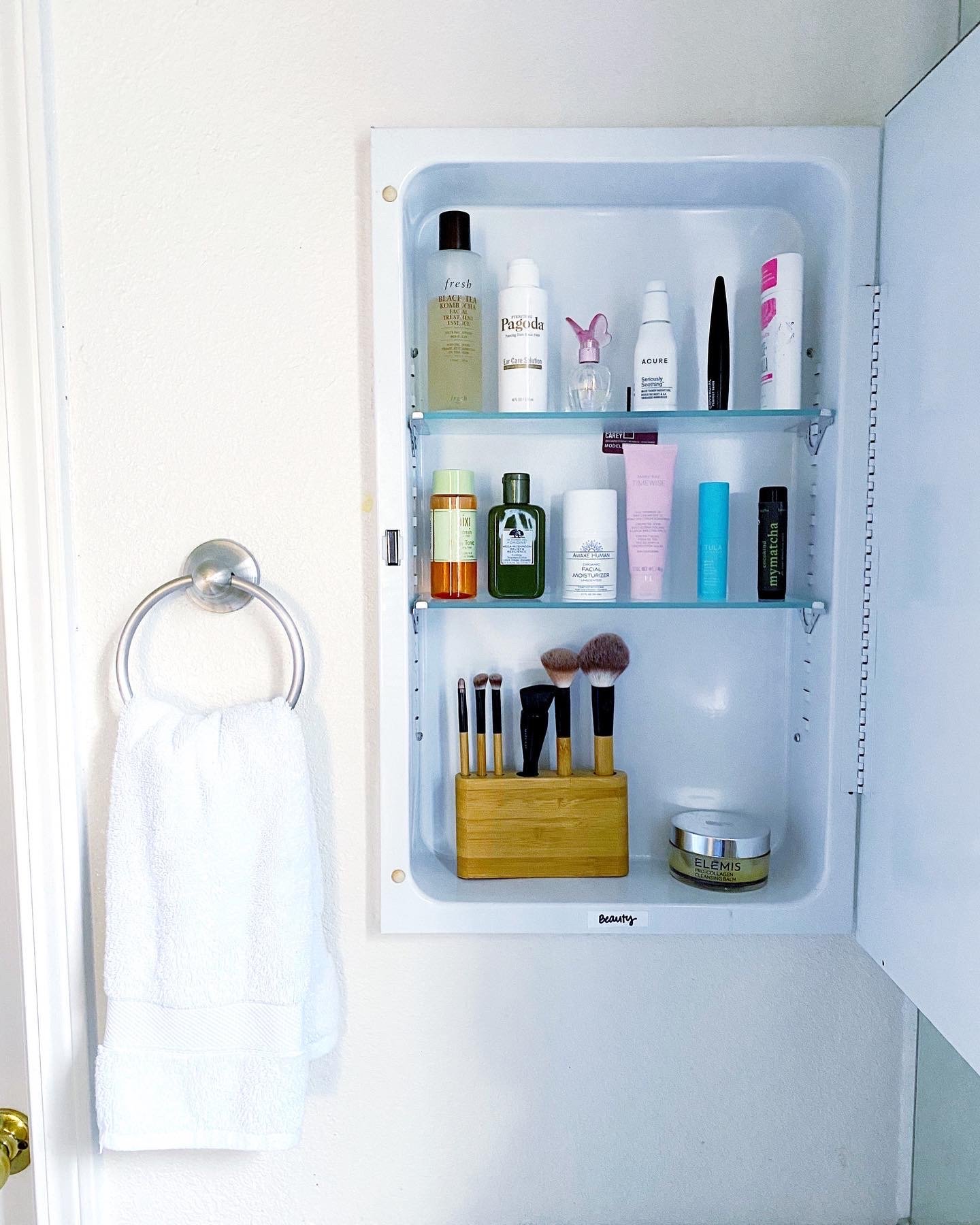 Bathroom cabinet filled with skincare and makeup products, brushes in a wooden holder, and a white towel hanging on a silver towel ring