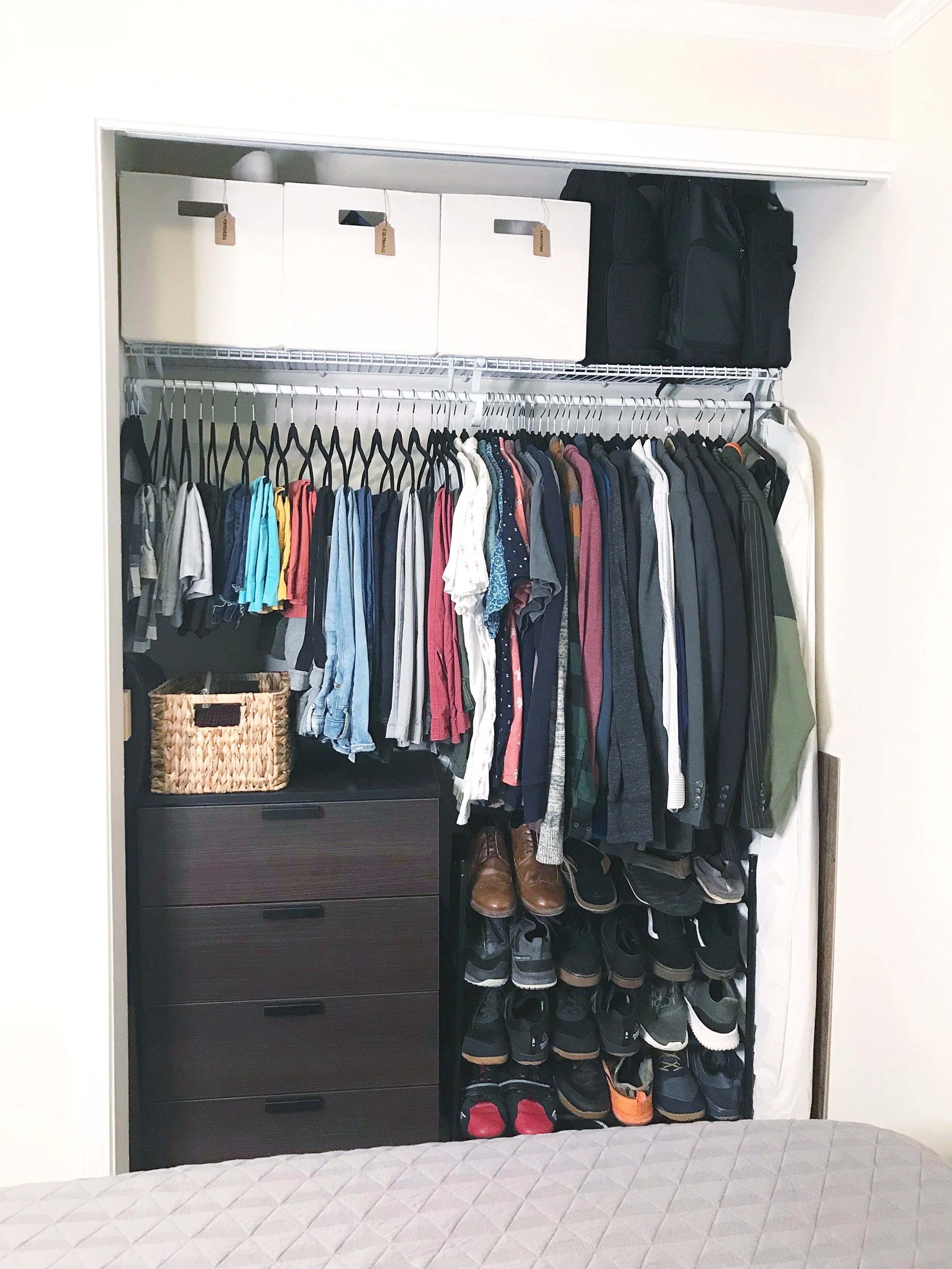 Open closet with hanging clothes, shoes on bottom, storage drawers, boxes, and bags on top shelf.