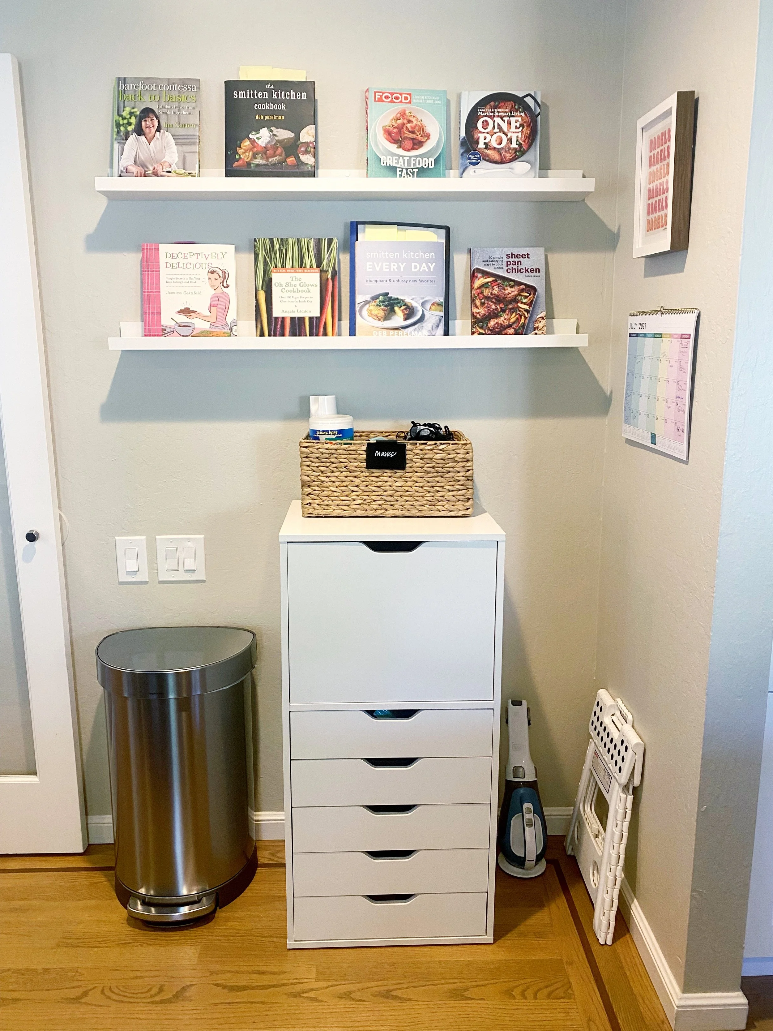 Room corner with two white shelves filled with cookbooks, a white storage cabinet, and various household items including a trash can, oven mitts, and an iron. A 2021 July calendar is on the wall.