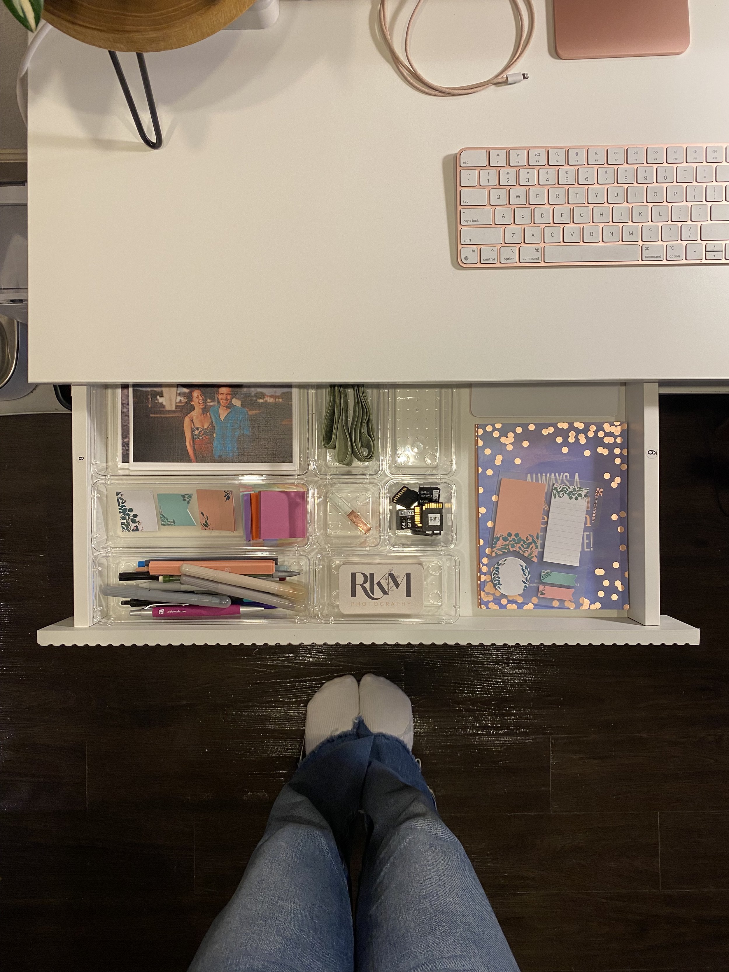 Top-down view of a white desk with a pink keyboard, a pair of pink headphones, a pink phone, and a small pink notebook. Below the desk, a person wearing jeans and white socks is standing on a dark wooden floor, with an open drawer containing pens, st