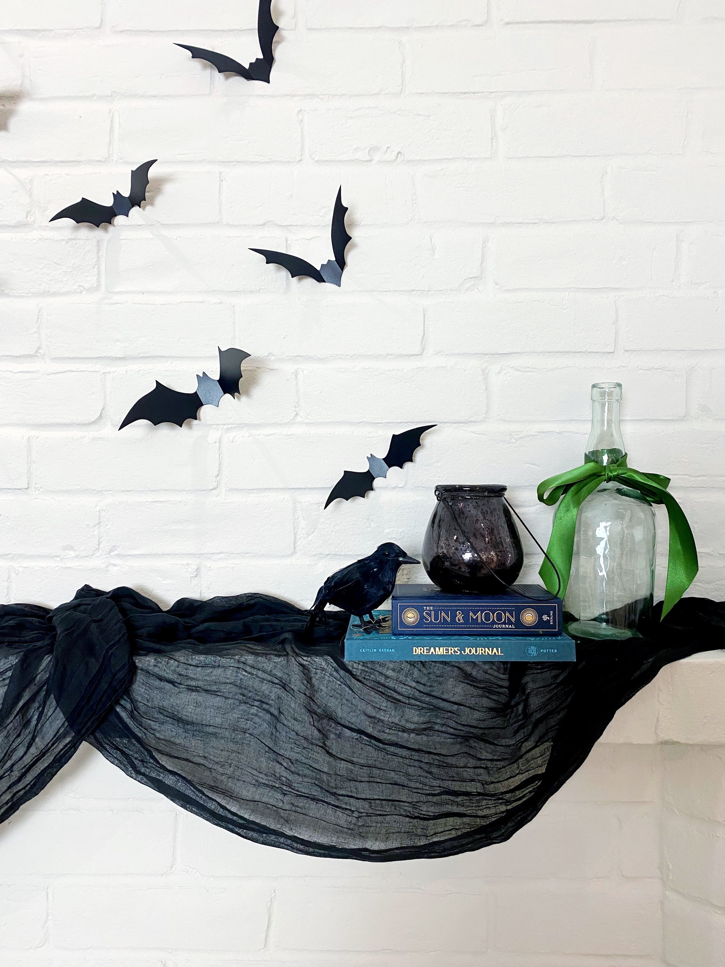 Halloween-themed decor featuring black bats on the wall, a black crow figurine, two books titled 'Sun & Moon' and 'Dreamer's Journal,' a black jar, and a glass bottle wrapped with a green ribbon, all arranged on a black fabric draped over a white bri
