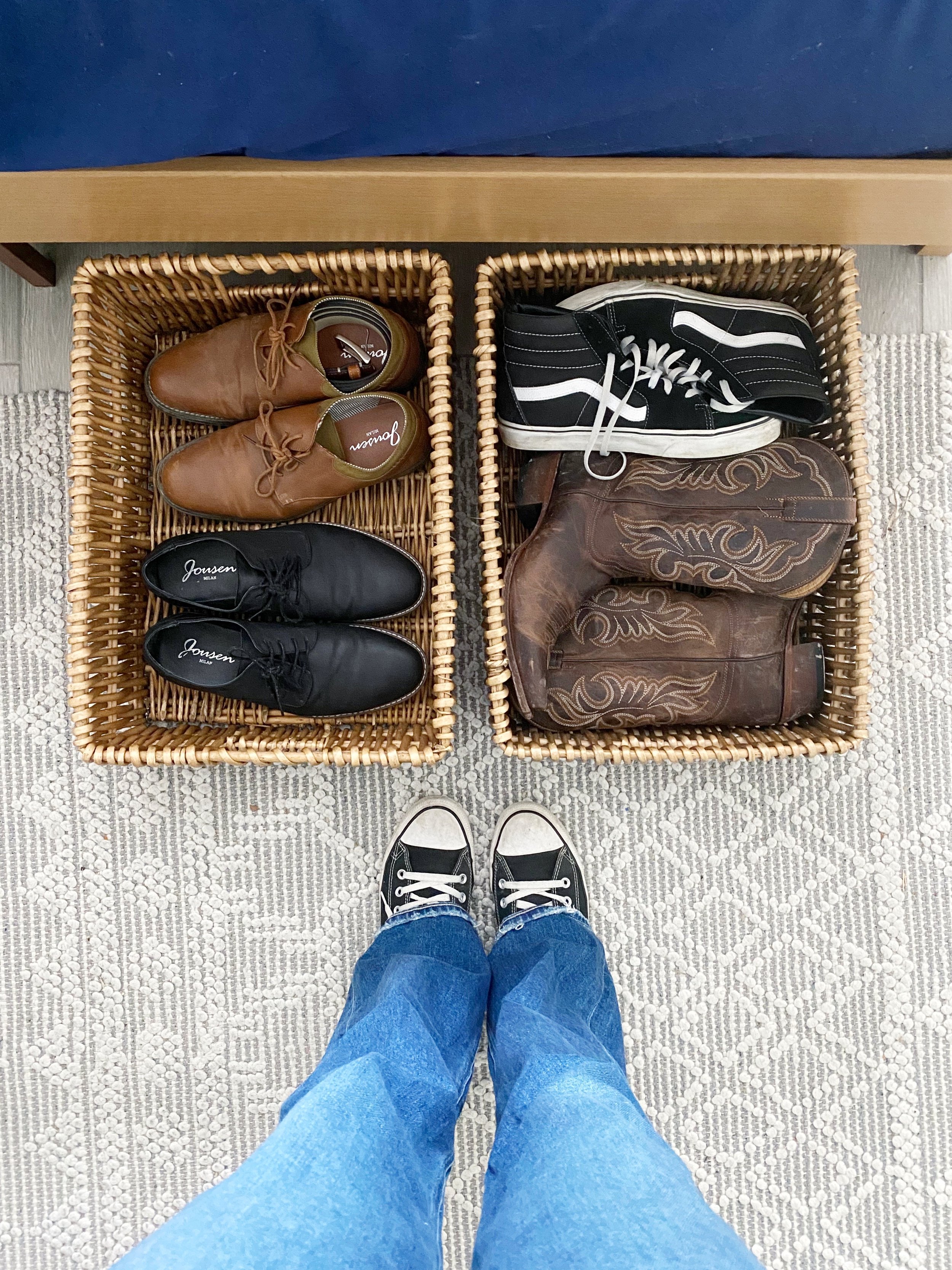 Person wearing black and white sneakers standing in front of two wicker baskets filled with various shoes and boots, on a patterned rug.